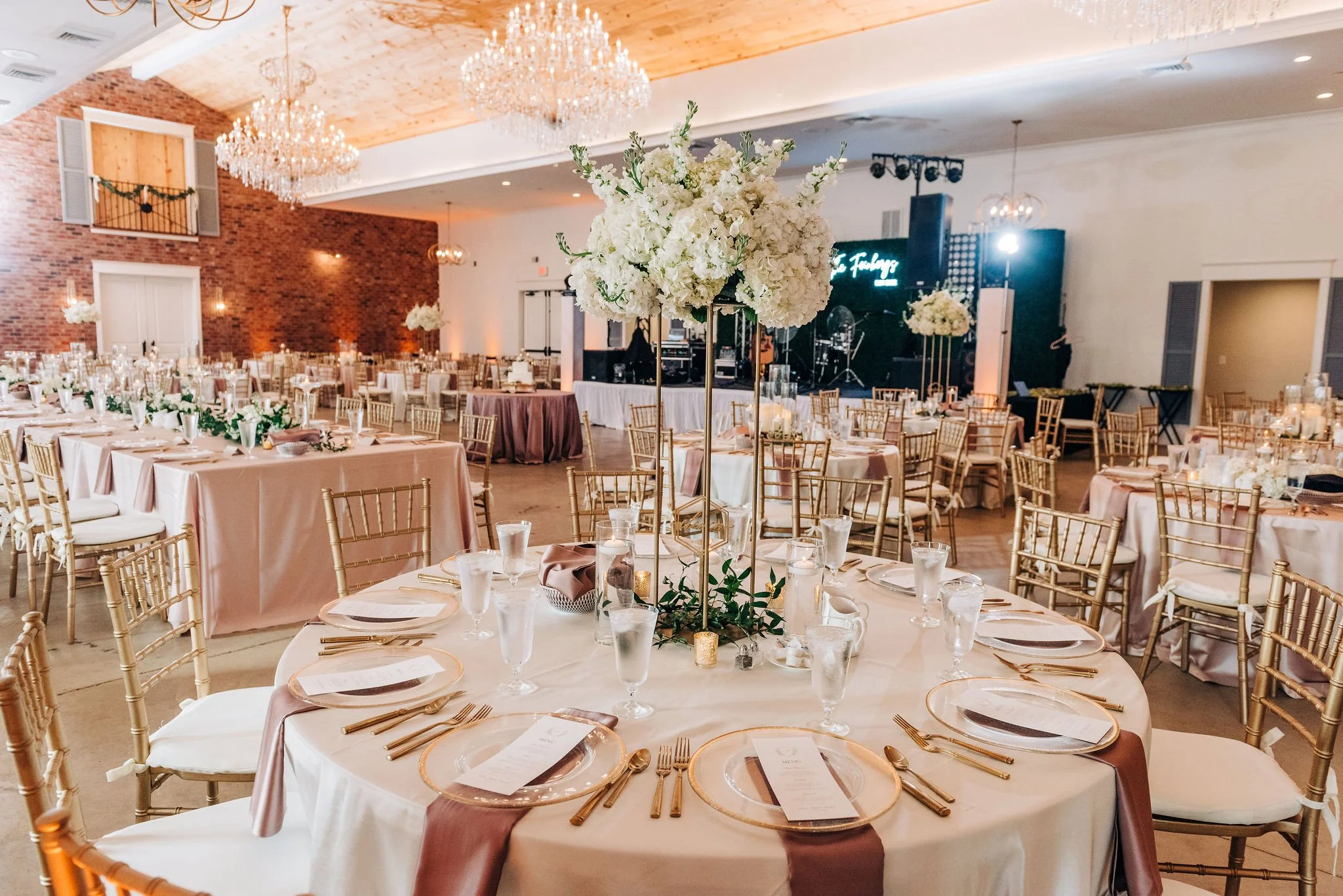 Elegant event hall decorated with long tables covered in blush pink tablecloths, gold chairs, floral centerpieces, and candles. Stage with musical instruments and signage in the background, chandeliers hanging from a wooden ceiling, and exposed brick walls.