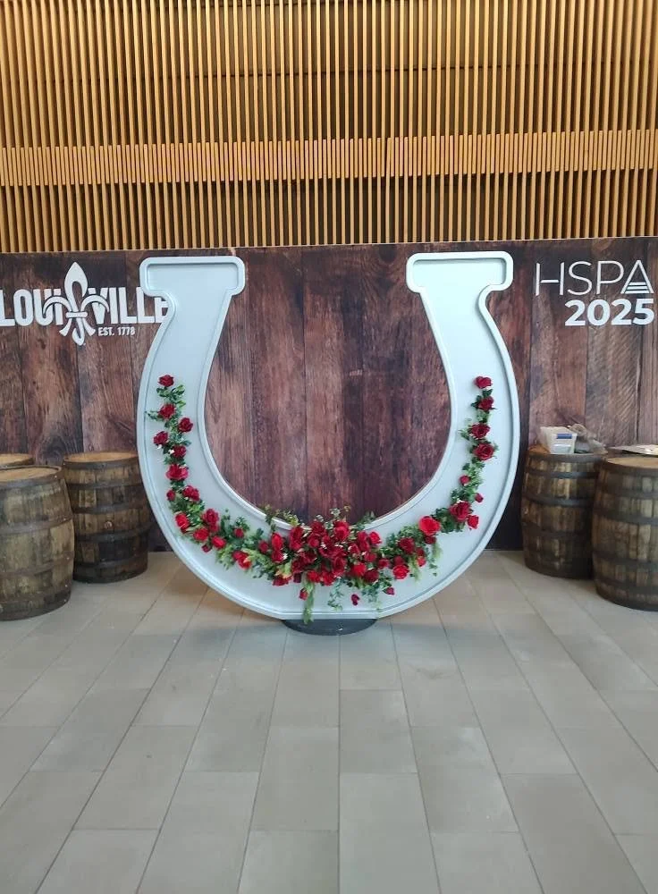 Large white horseshoe-shaped decorative piece decorated with red and green floral arrangement, flanked by wooden barrels on both sides, with a wooden wall and vertical slats in the background.