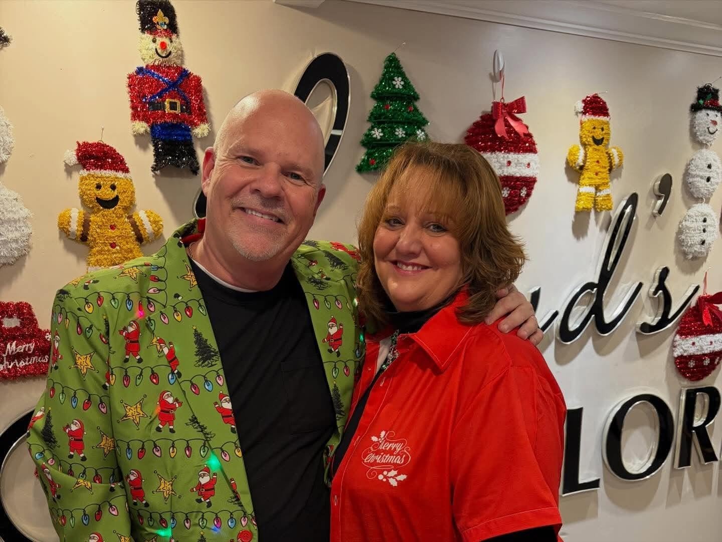 A smiling man and woman pose in front of a decorated wall with Christmas ornaments, including a Christmas tree, gingerbread men, snowmen, and Santa Claus figures, in a festive indoor setting.