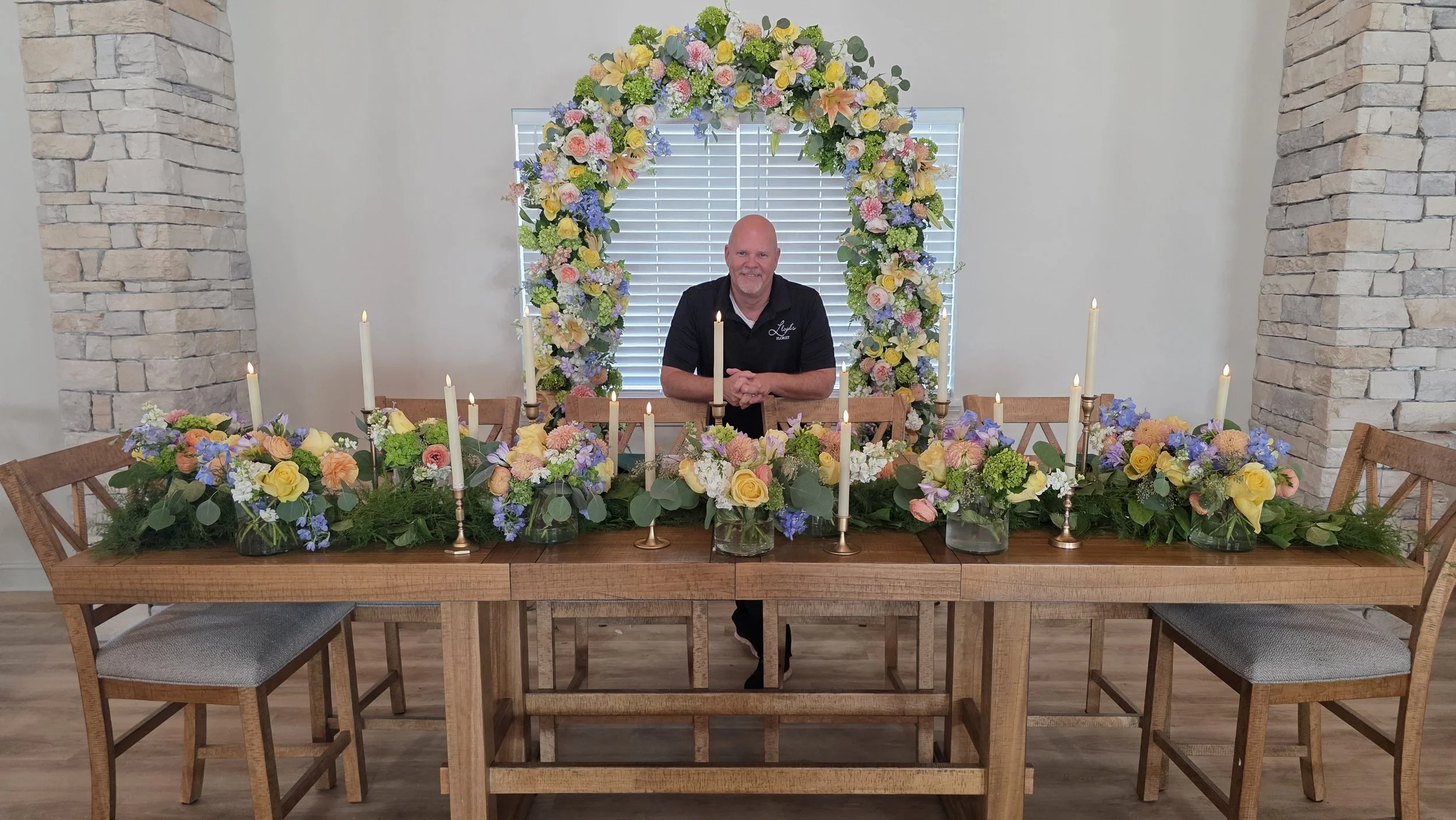 A man sitting at a decorated dining table with multiple floral arrangements and candle holders, and a floral arch behind him in a bright room with white walls and window blinds.