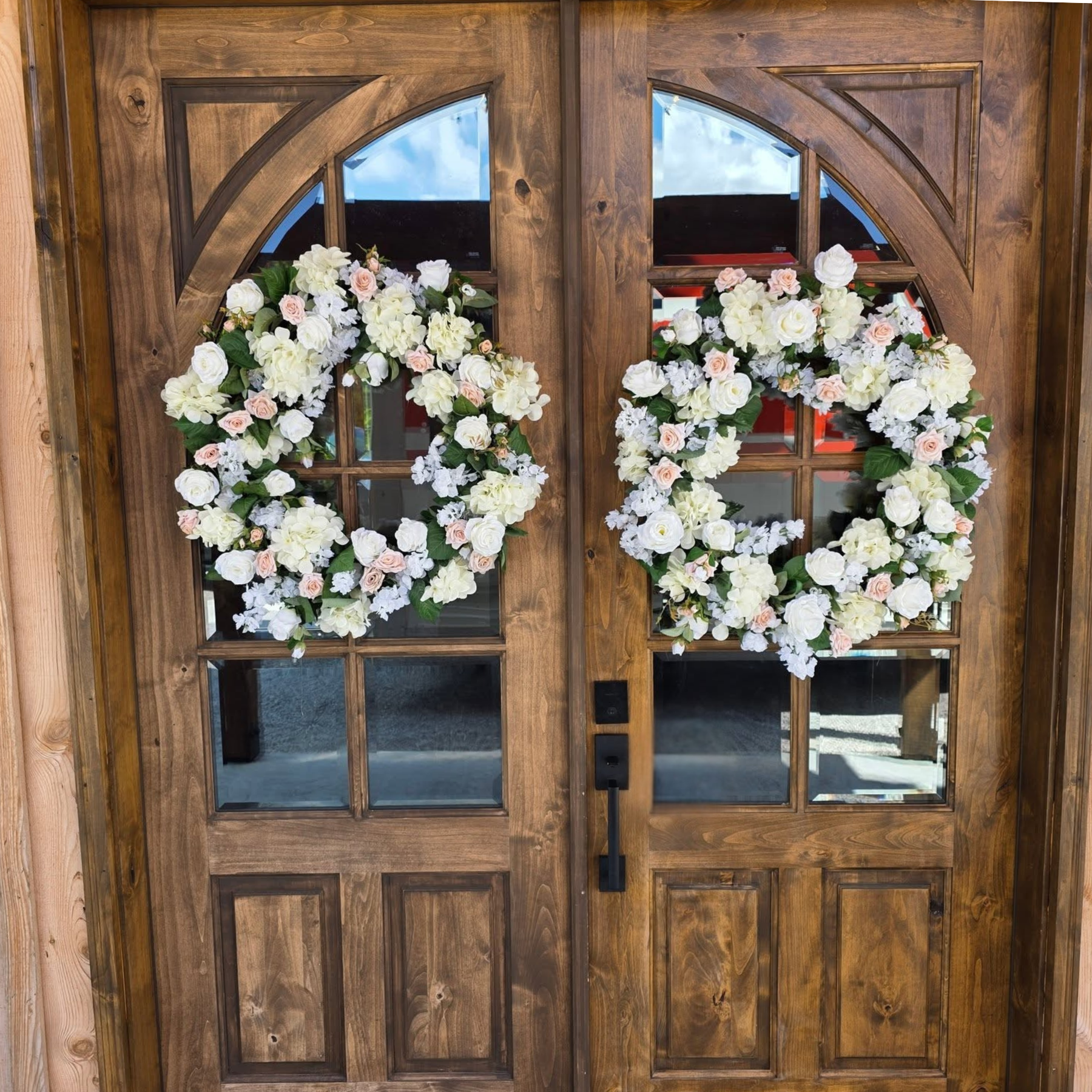 A wooden front door with multiple glass panels, decorated with two large floral wreaths made of white and pale pink roses and greenery.