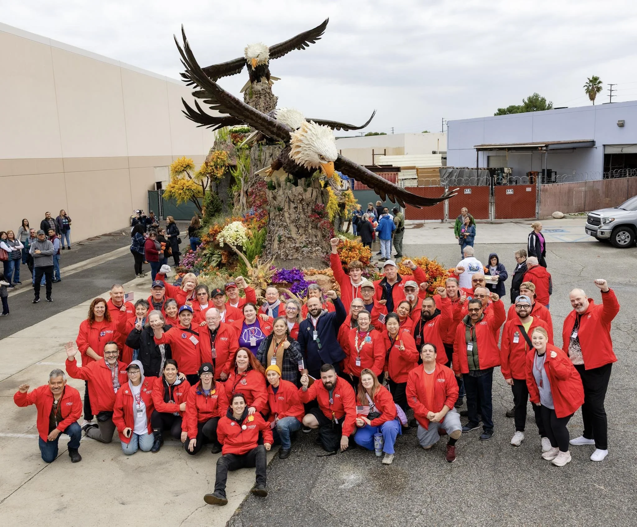 A large group of people wearing red jackets gathered in front of a monument of two eagles with outstretched wings, situated on a rock surrounded by colorful flowers. The scene appears to be festive or commemorative, with some individuals smiling and raising their fists.