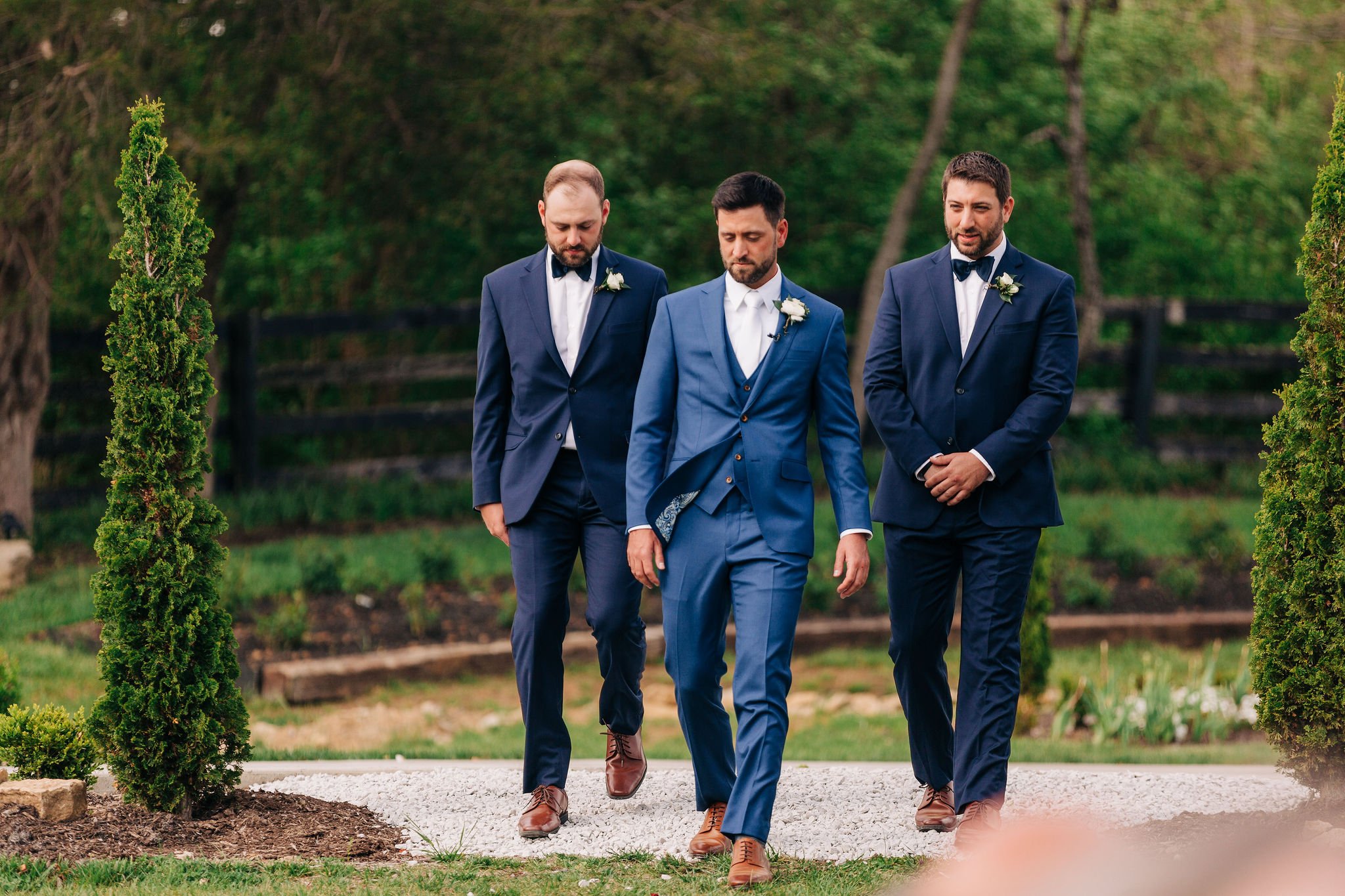 Three men in formal suits walking outdoors, possibly at a wedding or special event, surrounded by greenery and trees.