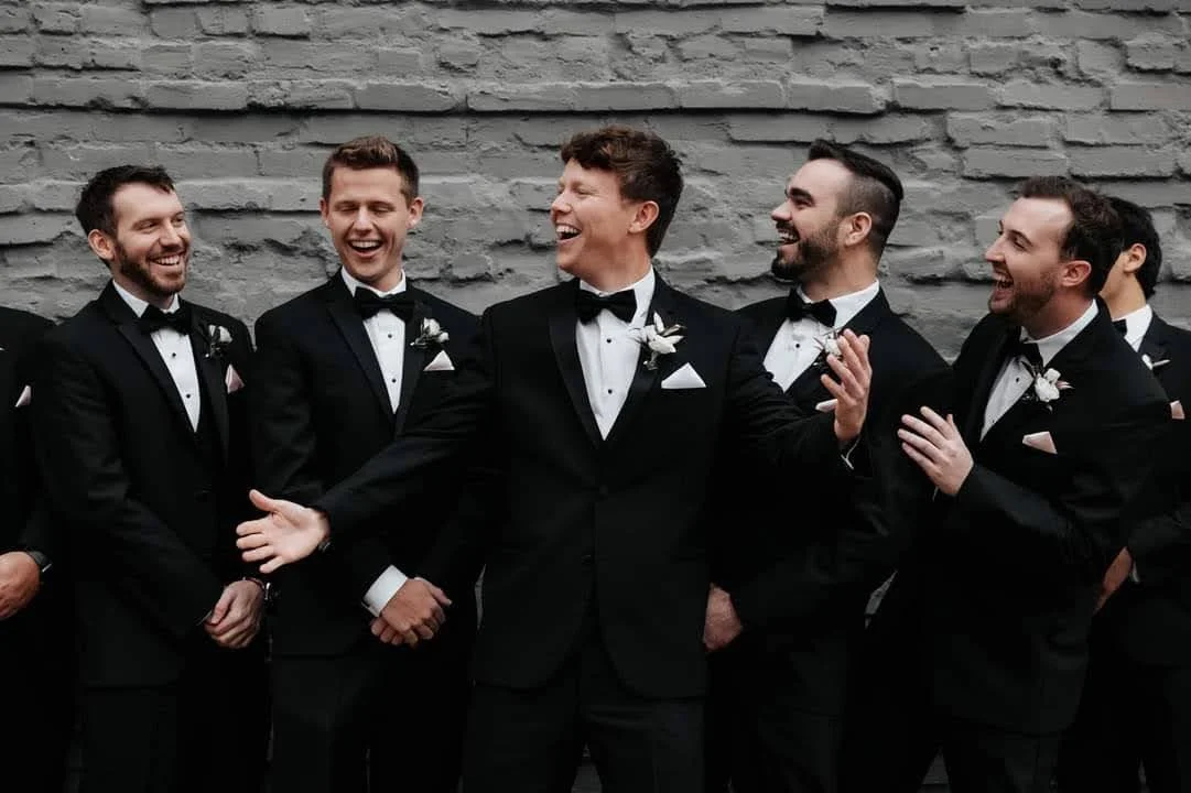 A group of men dressed in tuxedos and bow ties, smiling and laughing in front of a gray brick wall. They appear to be at a formal event or wedding.