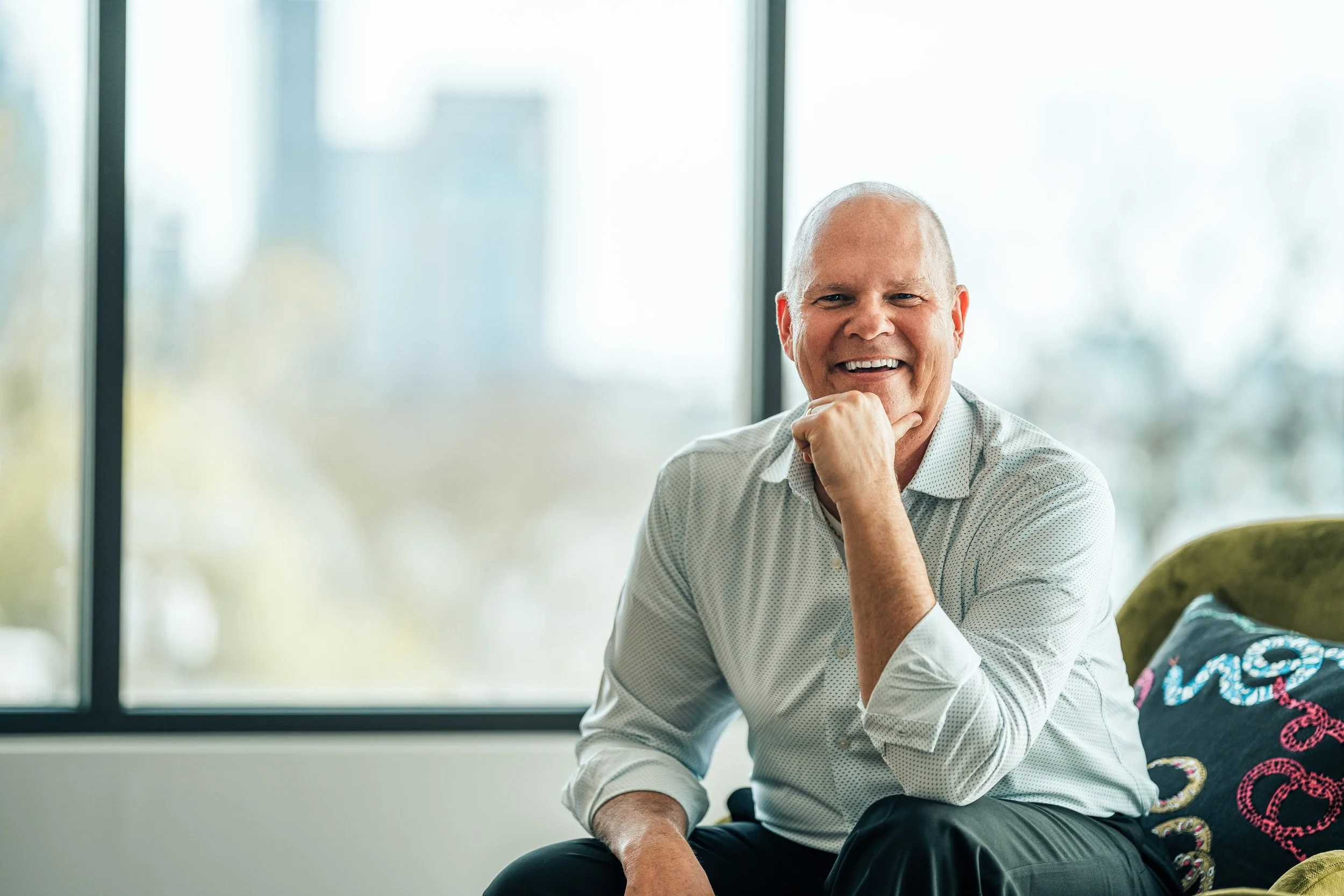 Smiling older man with white hair and a light shirt sitting indoors near large windows.