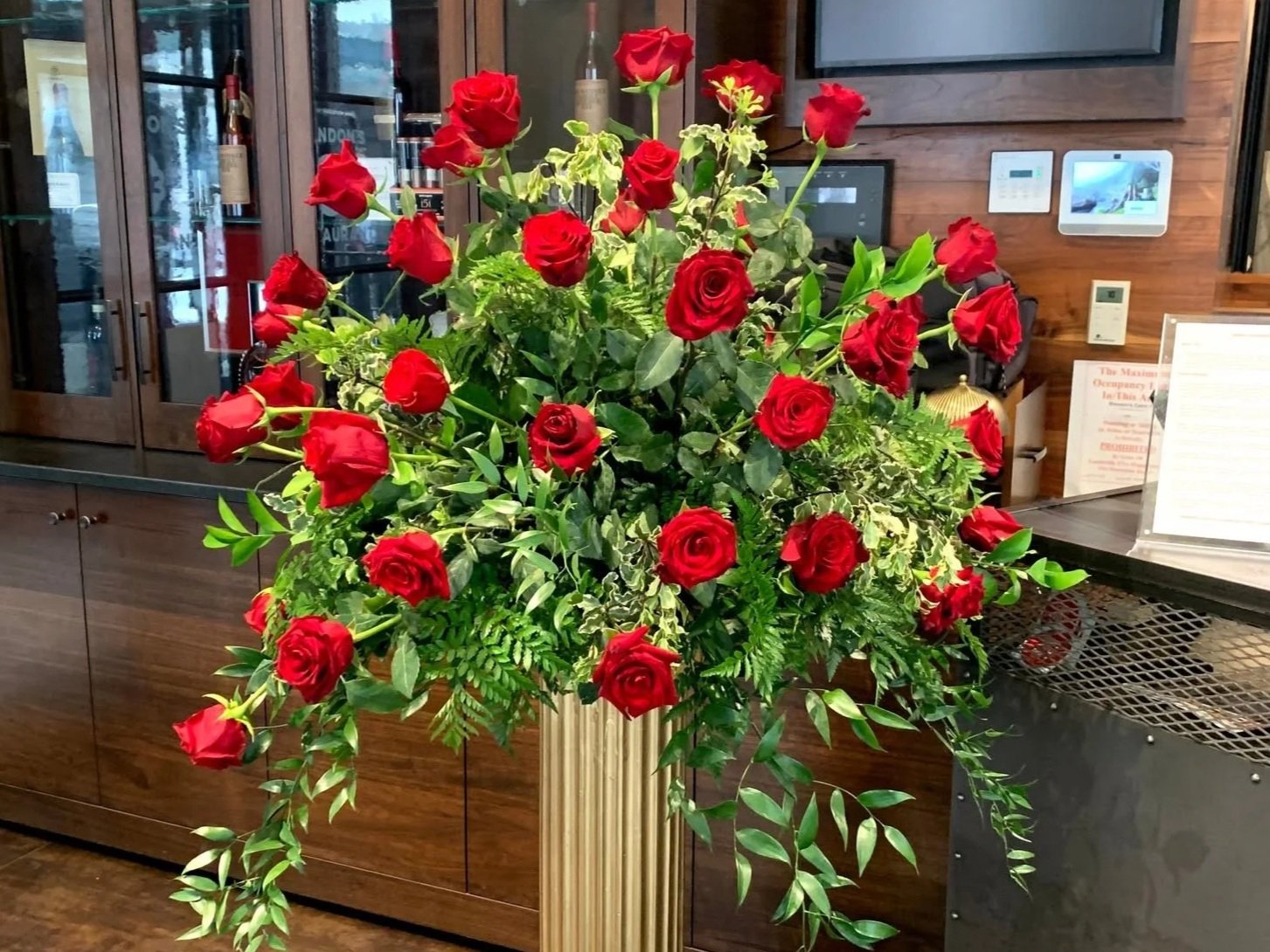 A large bouquet of red roses with green foliage in a tall gold vase on a wooden counter inside a restaurant or cafe.