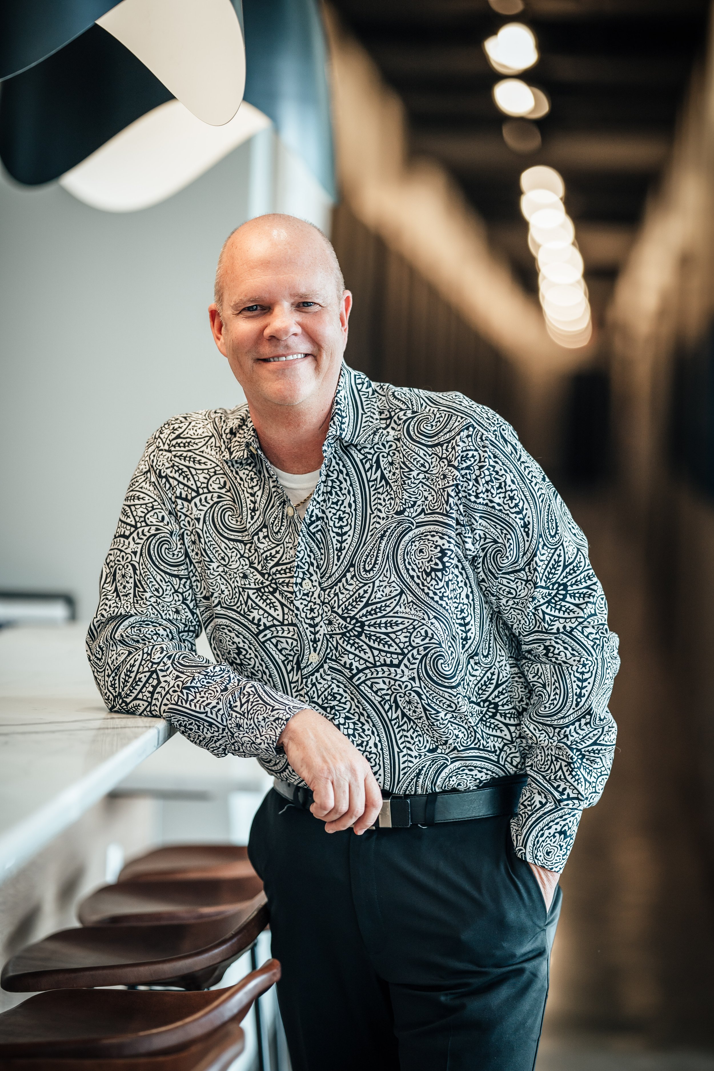 A man with a bald head and a big smile is standing in a modern indoor space with a blurred background of a corridor with overhead lighting. He is wearing a patterned black and white shirt and black pants, resting his right arm on a white counter with brown chairs nearby.