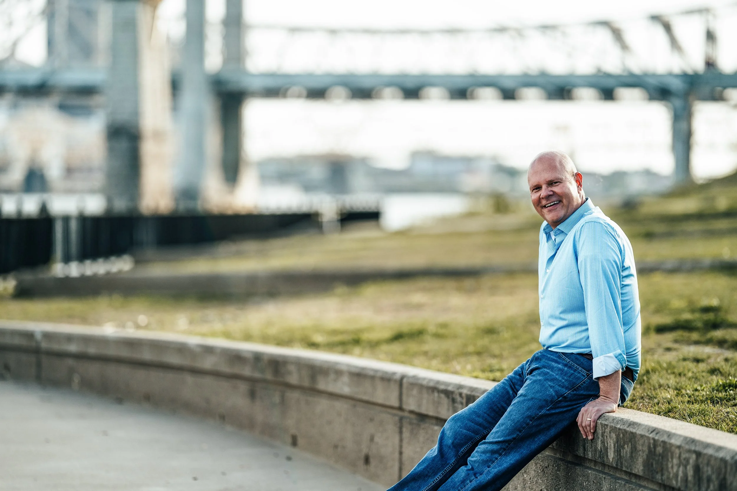 A bald man in a light blue shirt and jeans sitting on a concrete ledge in an outdoor setting with an industrial bridge structure in the background.