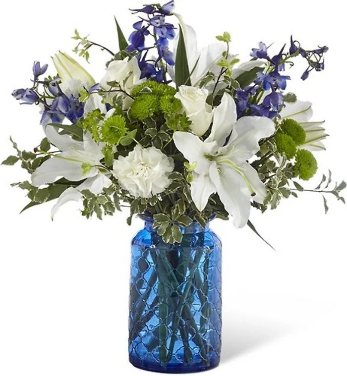A floral arrangement in a blue glass vase with white lilies, white roses, purple delphiniums, and green foliage.