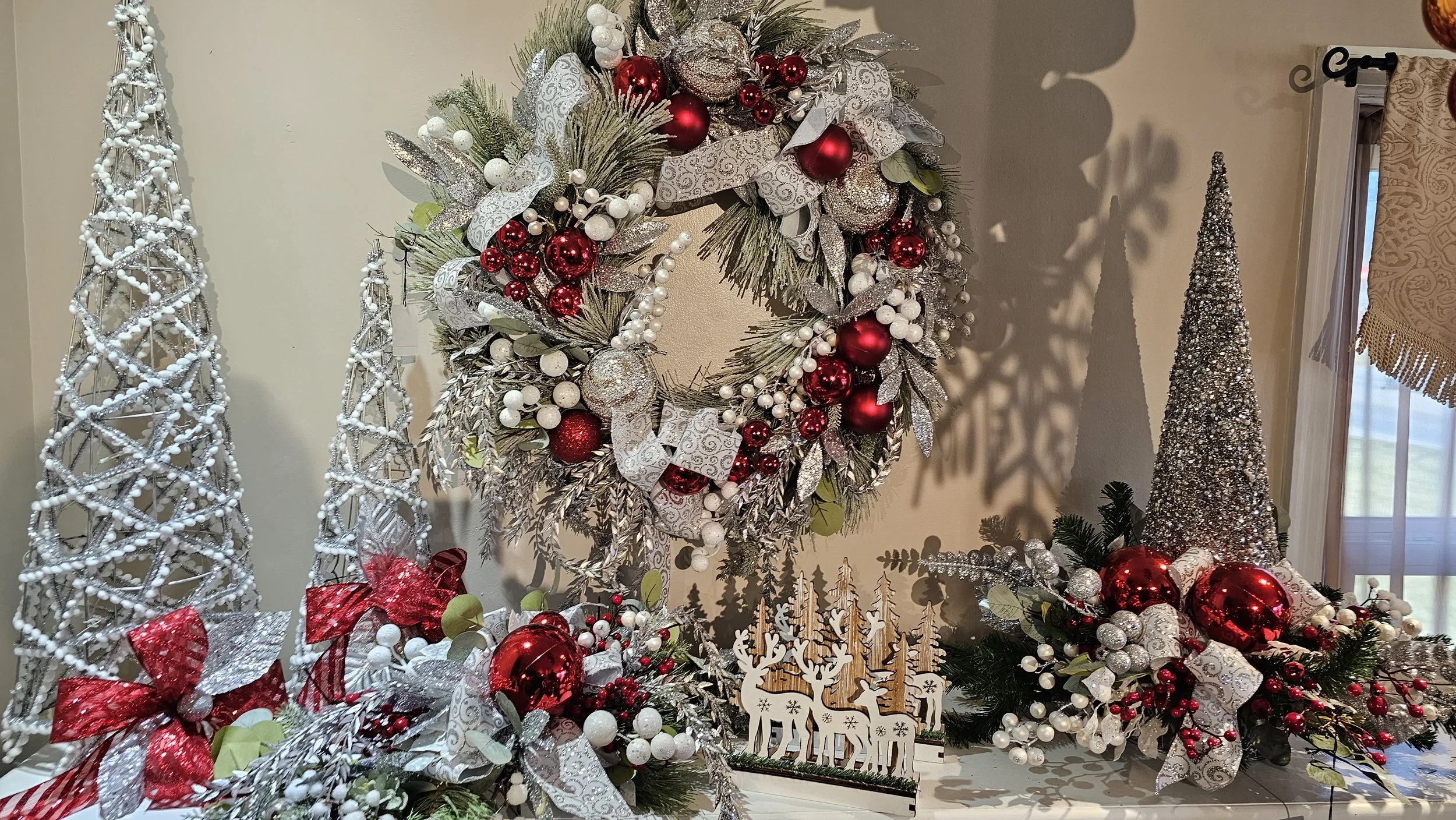 A Christmas wreath decorated with red and white ornaments, glittery leaves, and white ribbons, hanging on a wall. Surrounding decorations include tall white and silver cone-shaped Christmas trees and other festive ornaments.