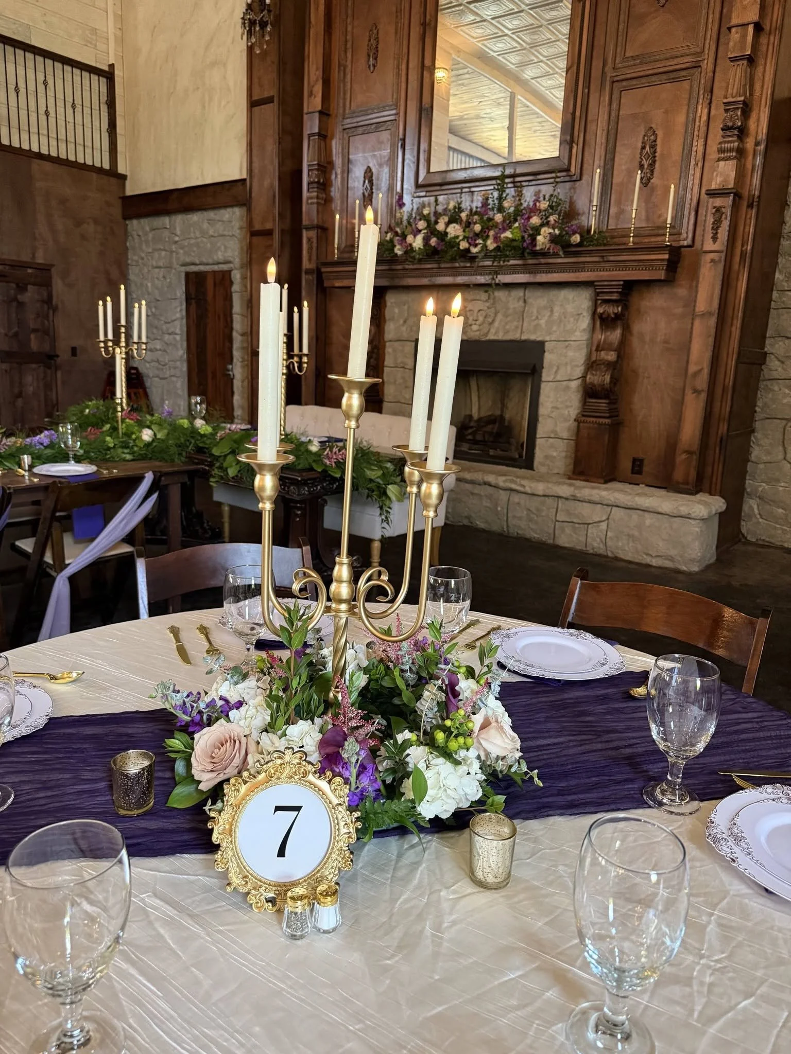 Elegant indoor event space with a table decorated with a floral centerpiece, tall white candles, and place settings. In the background, there is a stone fireplace with a large mirror above it and floral arrangements. Crystal candle holders and a table number 7 are also present.