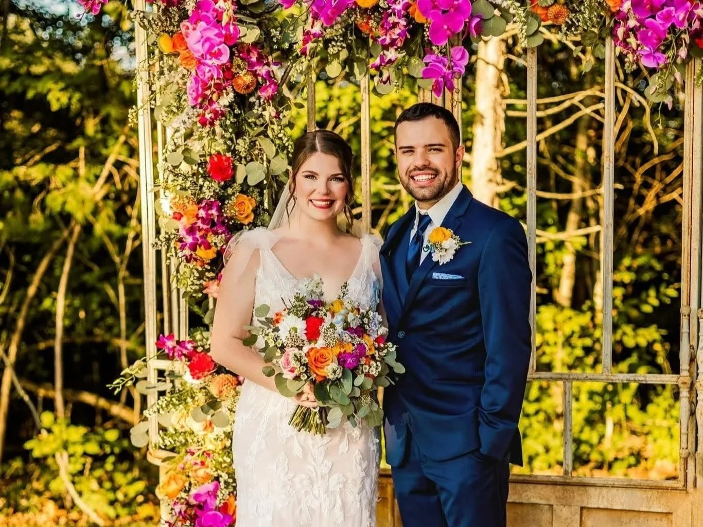 A bride and groom smiling outdoors at their wedding, standing in front of a metal arch decorated with colorful flowers, trees, and greenery.