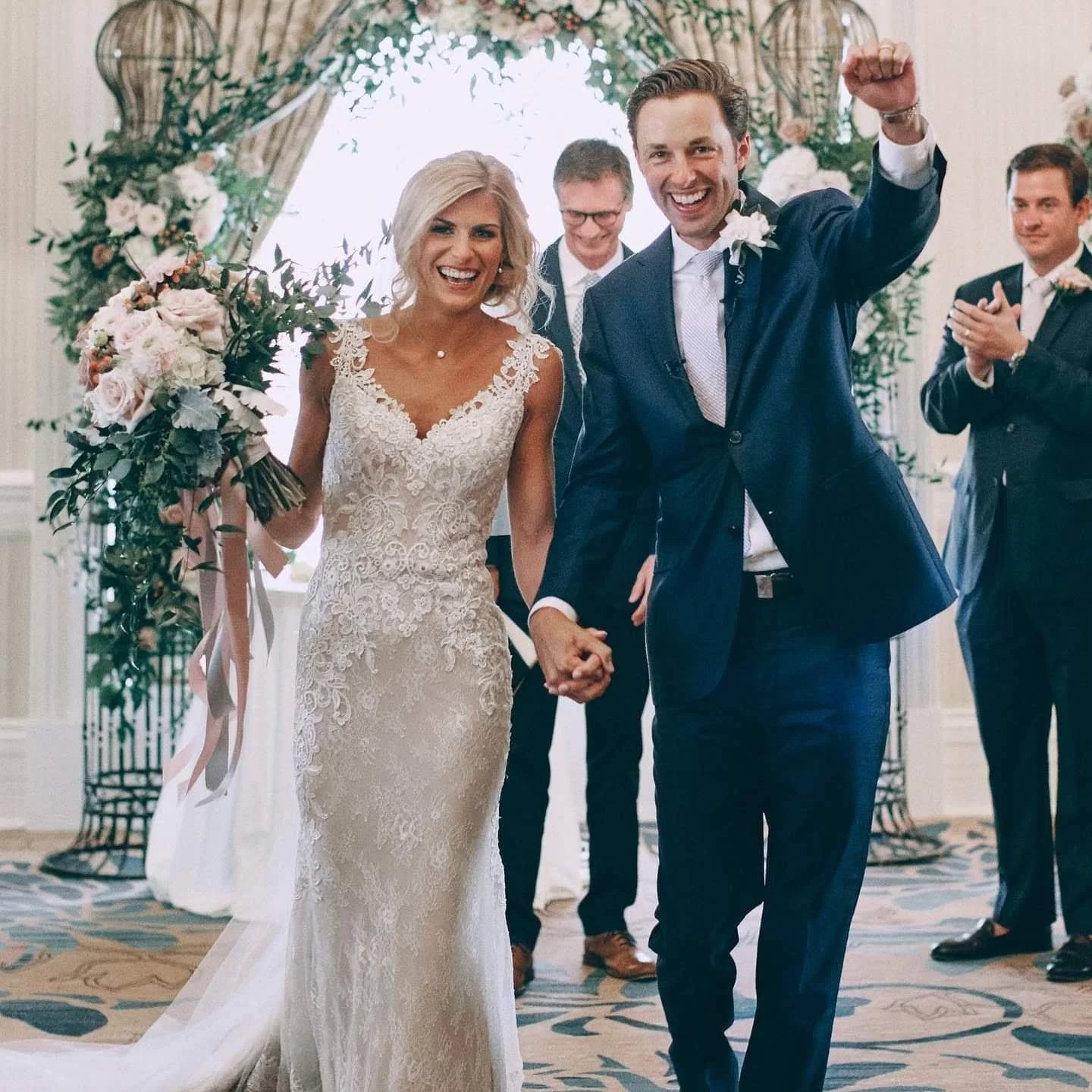 A newlywed couple holding hands and smiling, celebrating their wedding, surrounded by friends and family in a decorated indoor venue.