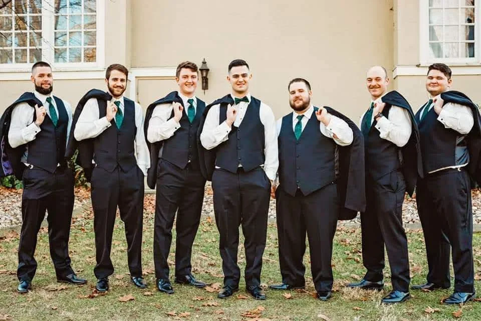 A group of eight men dressed in formal suits and tuxedos standing outdoors in front of a beige building with large windows. They are carrying their tuxedo jackets over their shoulders, smiling and posing for the photo.