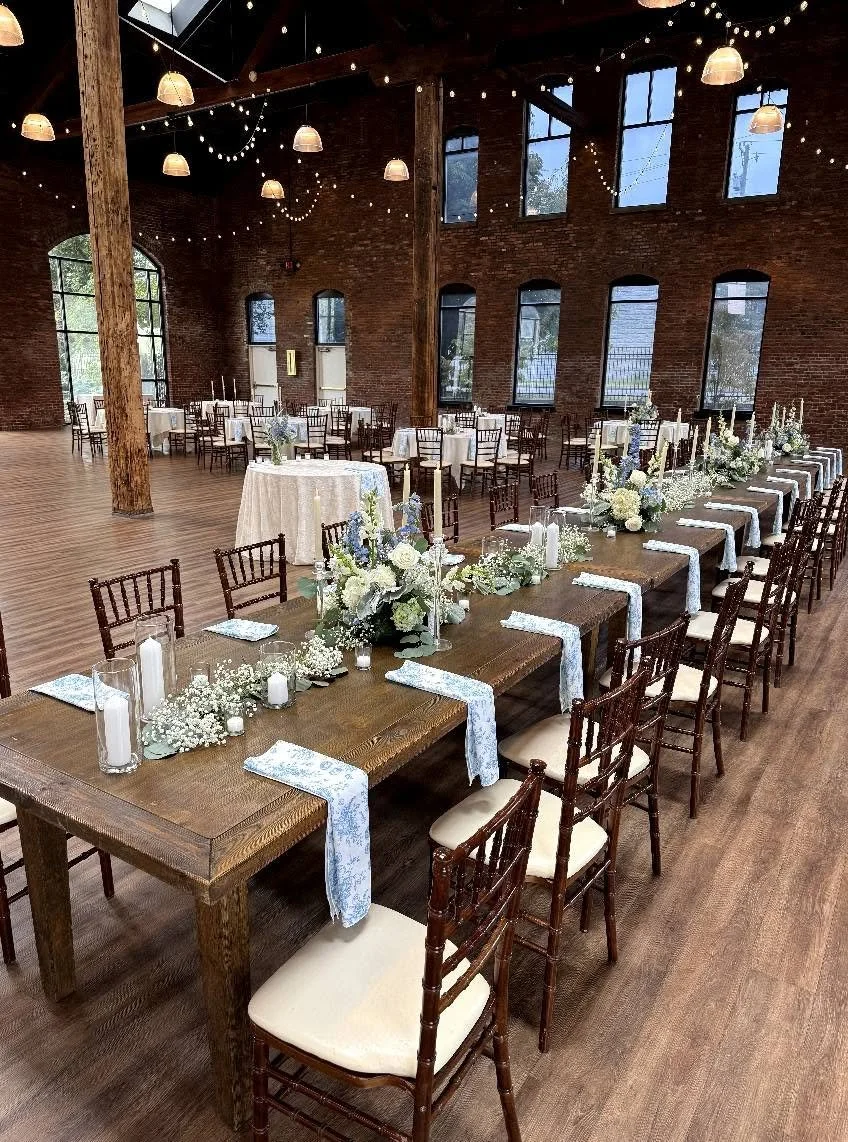 A decorated indoor event space with wooden floors and exposed brick walls. Long wooden tables with floral centerpieces, candles, and blue and white table runners are set up for a celebration. Additional round tables with chairs are arranged in the background. String lights and hanging lamps illuminate the space.