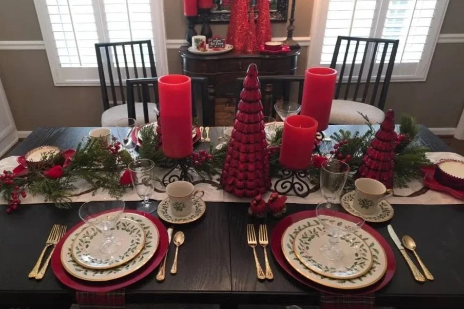 Festive holiday table set with red candles, Christmas themed plates, gold utensils, and greenery centerpiece.