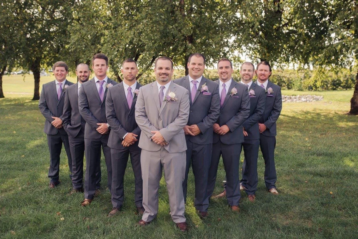 A group of nine men in suits, standing outdoors on grass with trees in the background, during a wedding or formal event. They are smiling and facing the camera.