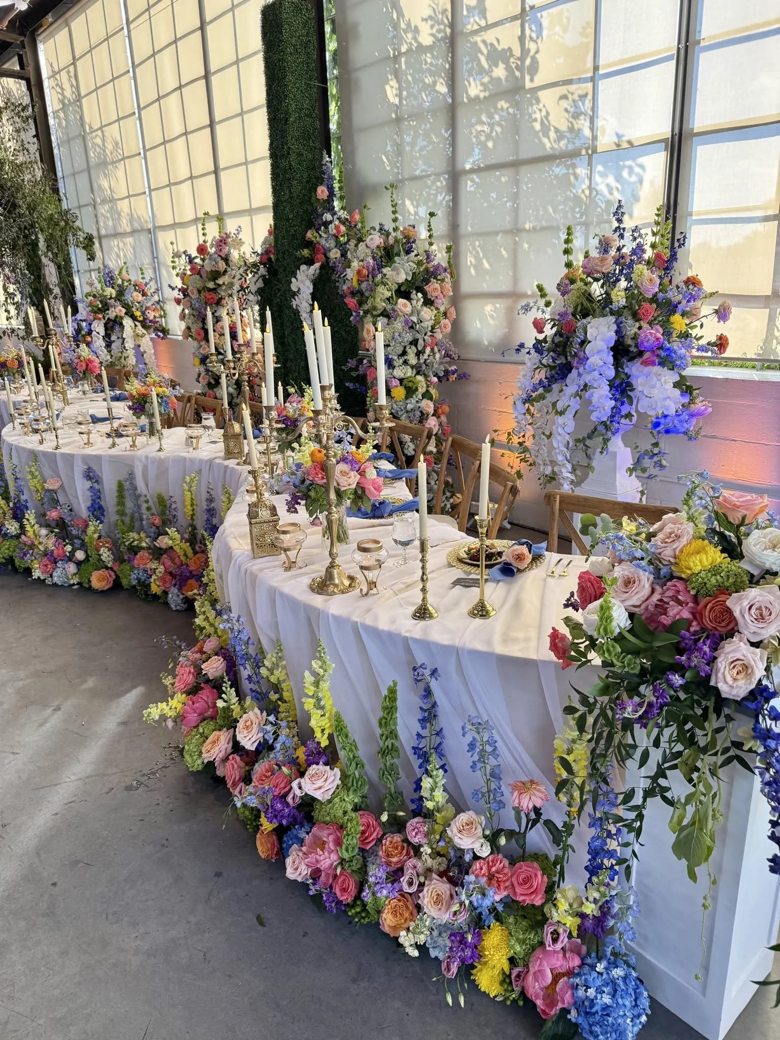 A long banquet table decorated with a white tablecloth and surrounded by chairs, adorned with colorful flower arrangements, candles, and tableware, set in a bright room with large windows and floral decorations on the wall.