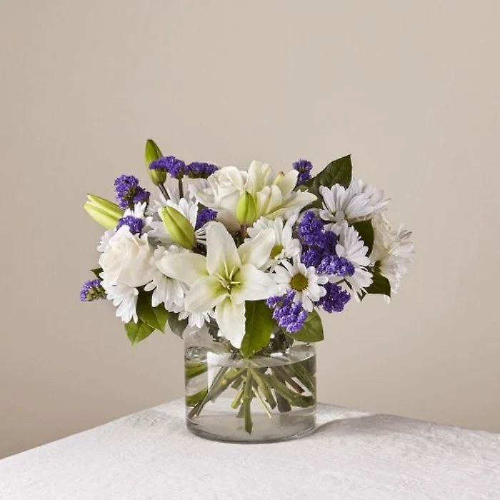 A bouquet of white lilies, daisies, and purple statice flowers in a glass vase on a white table.