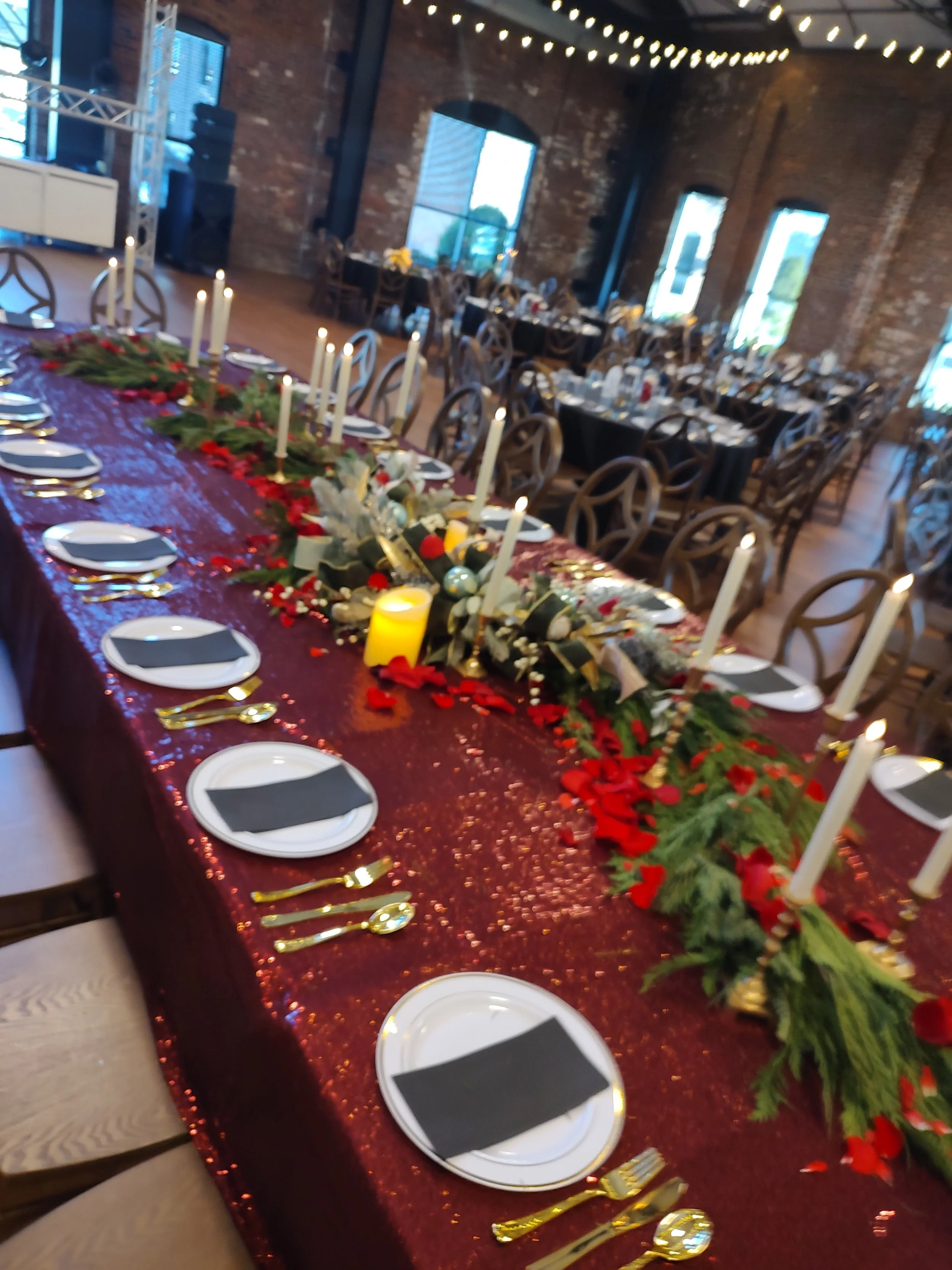 A long dining table decorated for a celebration, covered with a shiny red tablecloth, with white plates, gold utensils, black napkins, and tall candles in candle holders. The centerpiece is a festive arrangement of greenery, red flowers, and ornaments. In the background, there are multiple unoccupied chairs, exposed brick walls, large windows, and string lights hanging from the ceiling.