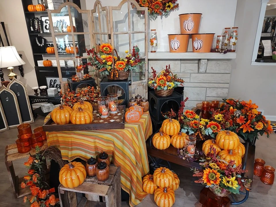 Fall-themed display with orange pumpkins, sunflower and autumn flower arrangements, candles, and seasonal decor in a cozy indoor setting.