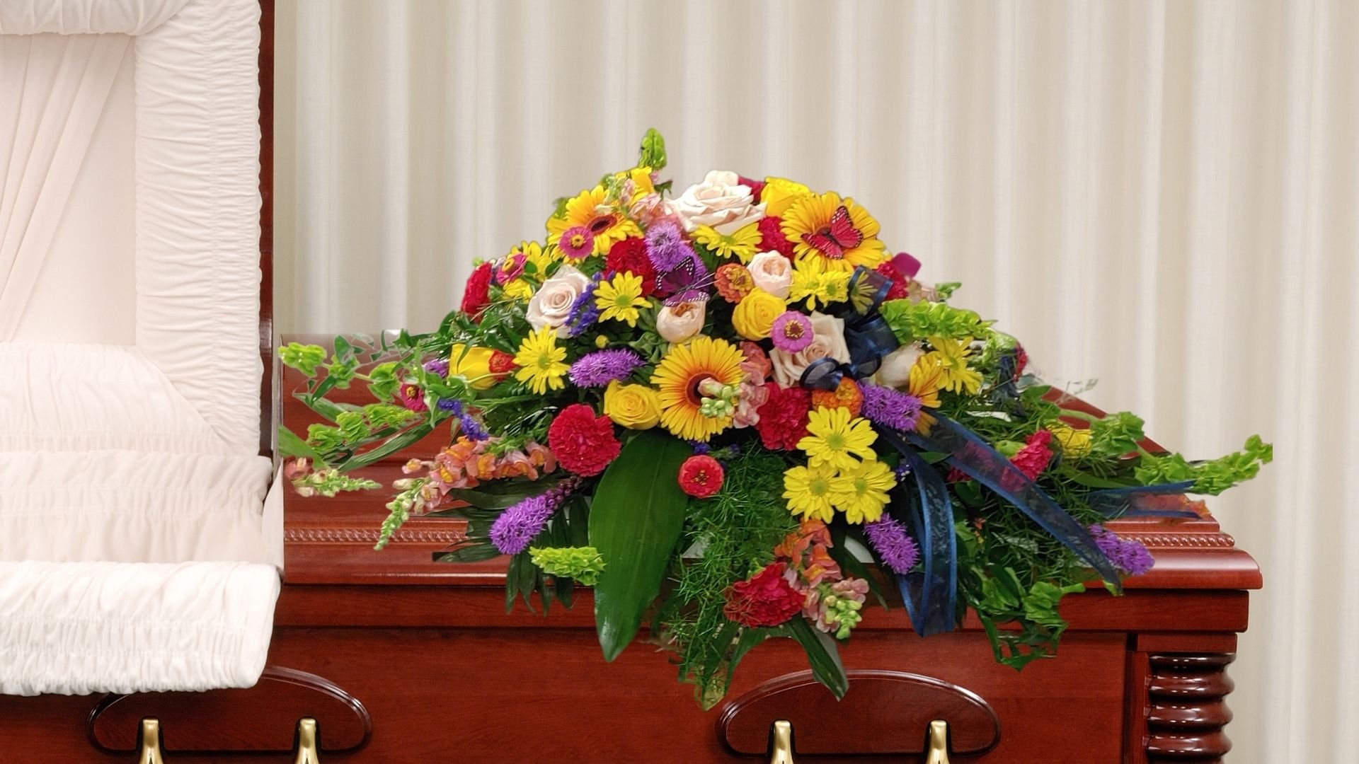 Colorful floral arrangement with yellow, pink, purple, and red flowers, placed on a wooden surface in front of a cream-colored curtain.