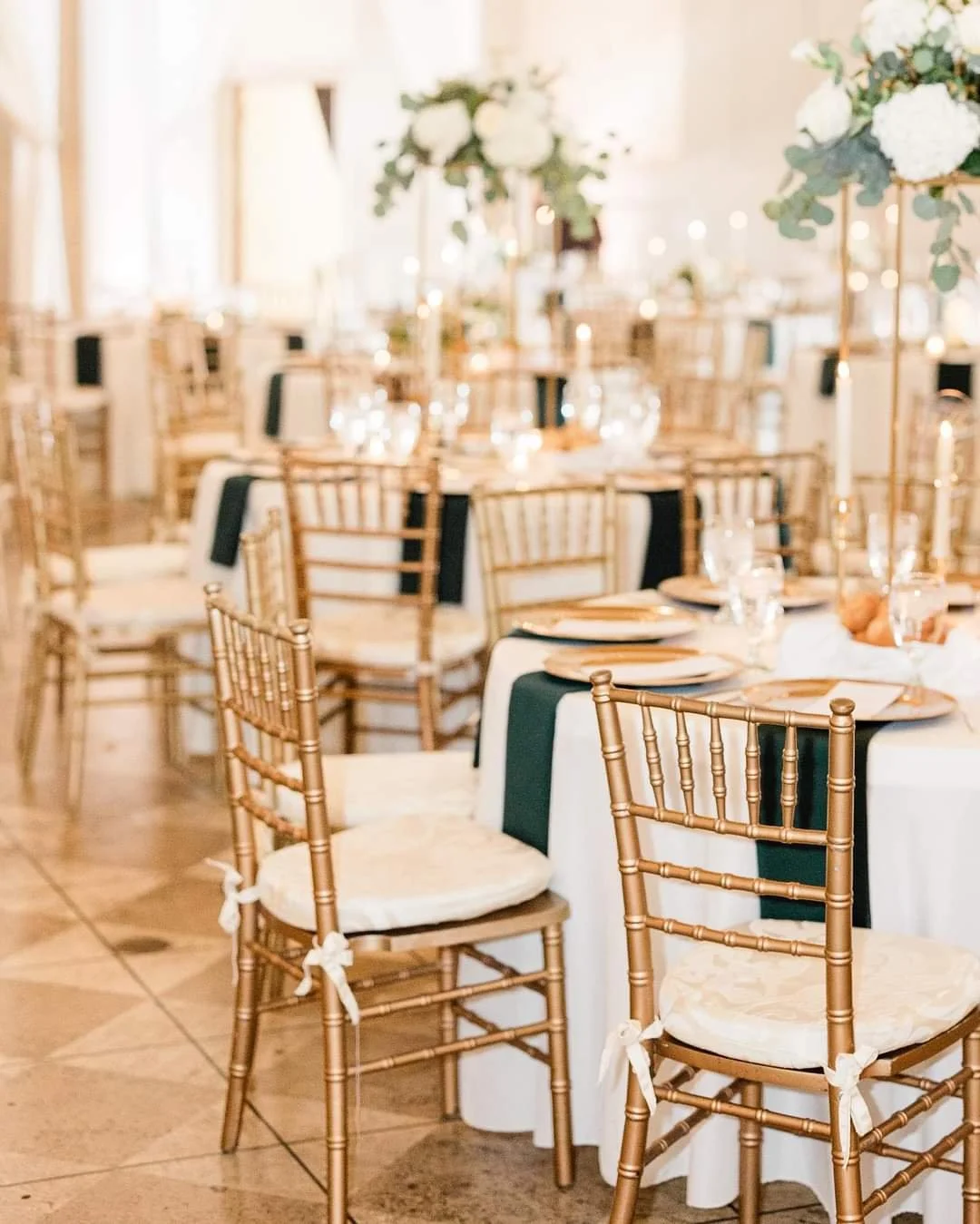 Elegant banquet hall decorated for a wedding reception with round tables, gold Chiavari chairs with white cushions, tall floral centerpieces, and candlelight.