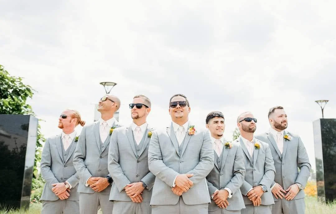 Group of seven men in light gray suits standing outdoors in a line, smiling and looking in different directions, with some wearing sunglasses and yellow boutonnières, likely at a wedding.