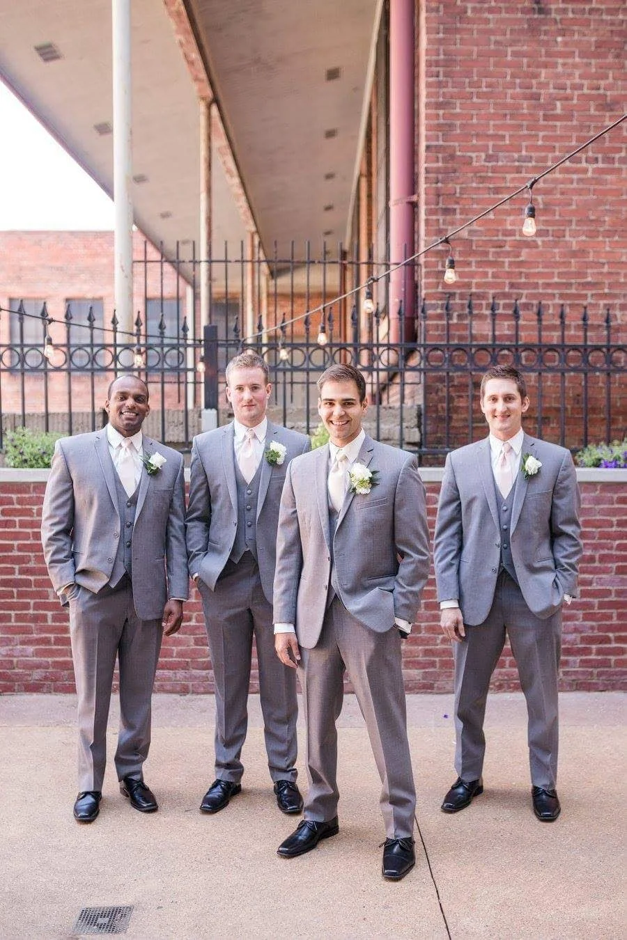 Four men dressed in gray suits and white shirts standing outdoors in front of a brick wall with string lights overhead.