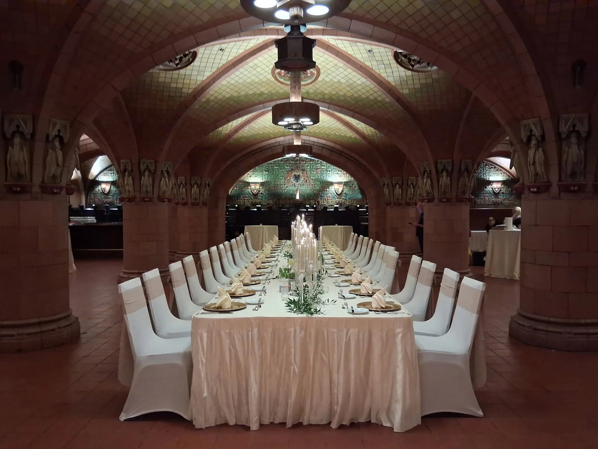 A long rectangular banquet table set for a formal dinner in an ornate dining hall with stone arches and decorative statues along the walls. The table is covered with a white tablecloth, set with wine glasses, plates, napkins, and floral centerpieces. The high ceiling has decorative lighting fixtures.