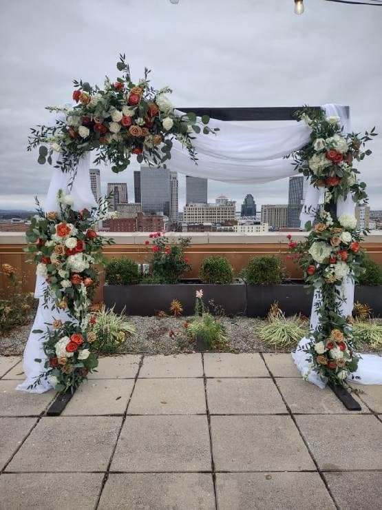 A floral wedding arch decorated with white and orange flowers and greenery, set on a rooftop with a city skyline in the background.