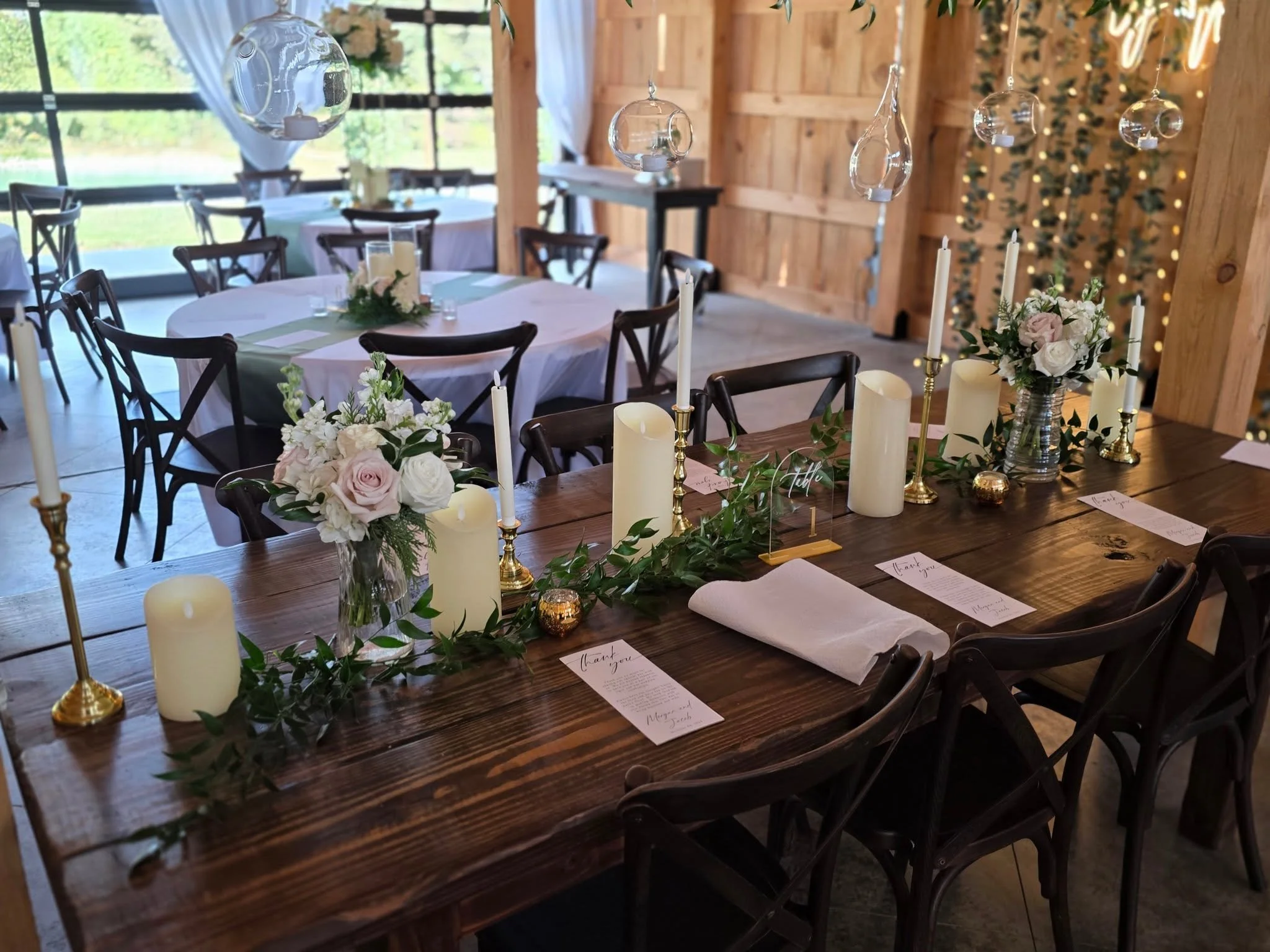 A rustic wooden indoor event space decorated with hanging glass globes, white candles, floral arrangements, and table settings for a celebration or wedding reception.