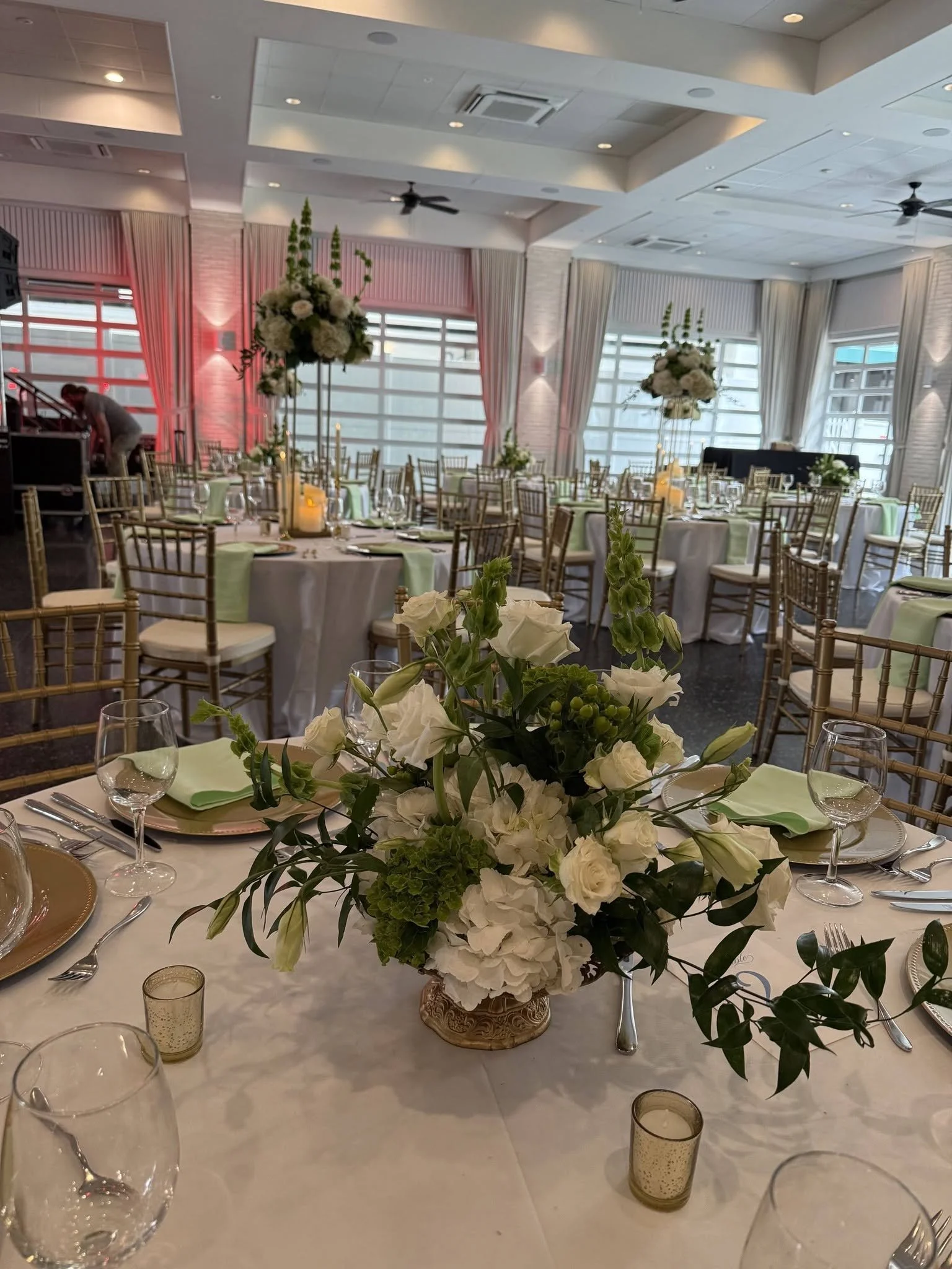 Elegant event reception table decorated with a flower centerpiece, with place settings, glasses, and candles, in a bright room with large windows and white curtains.