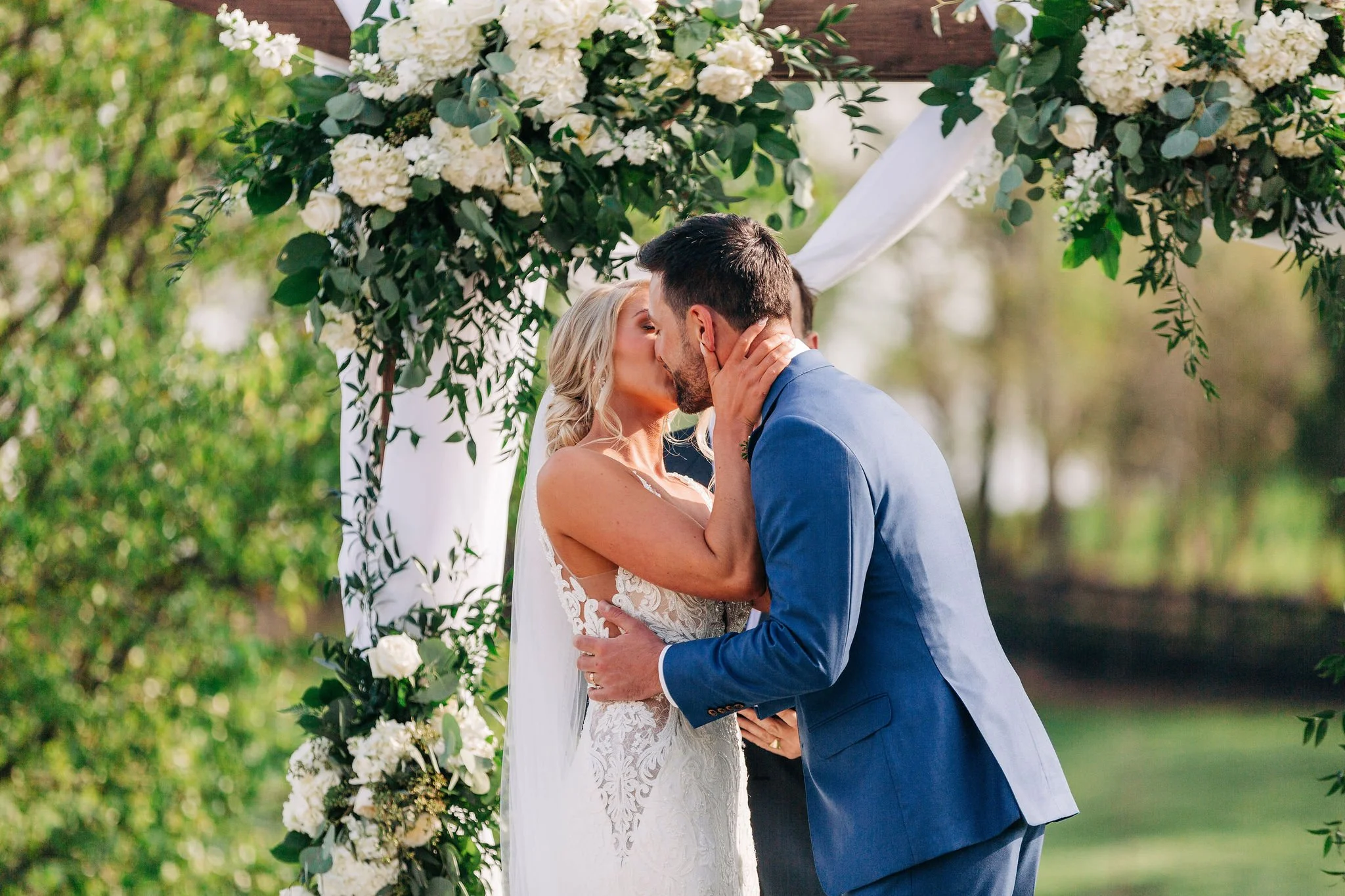 A bride and groom share a kiss during their outdoor wedding under a floral arch.
