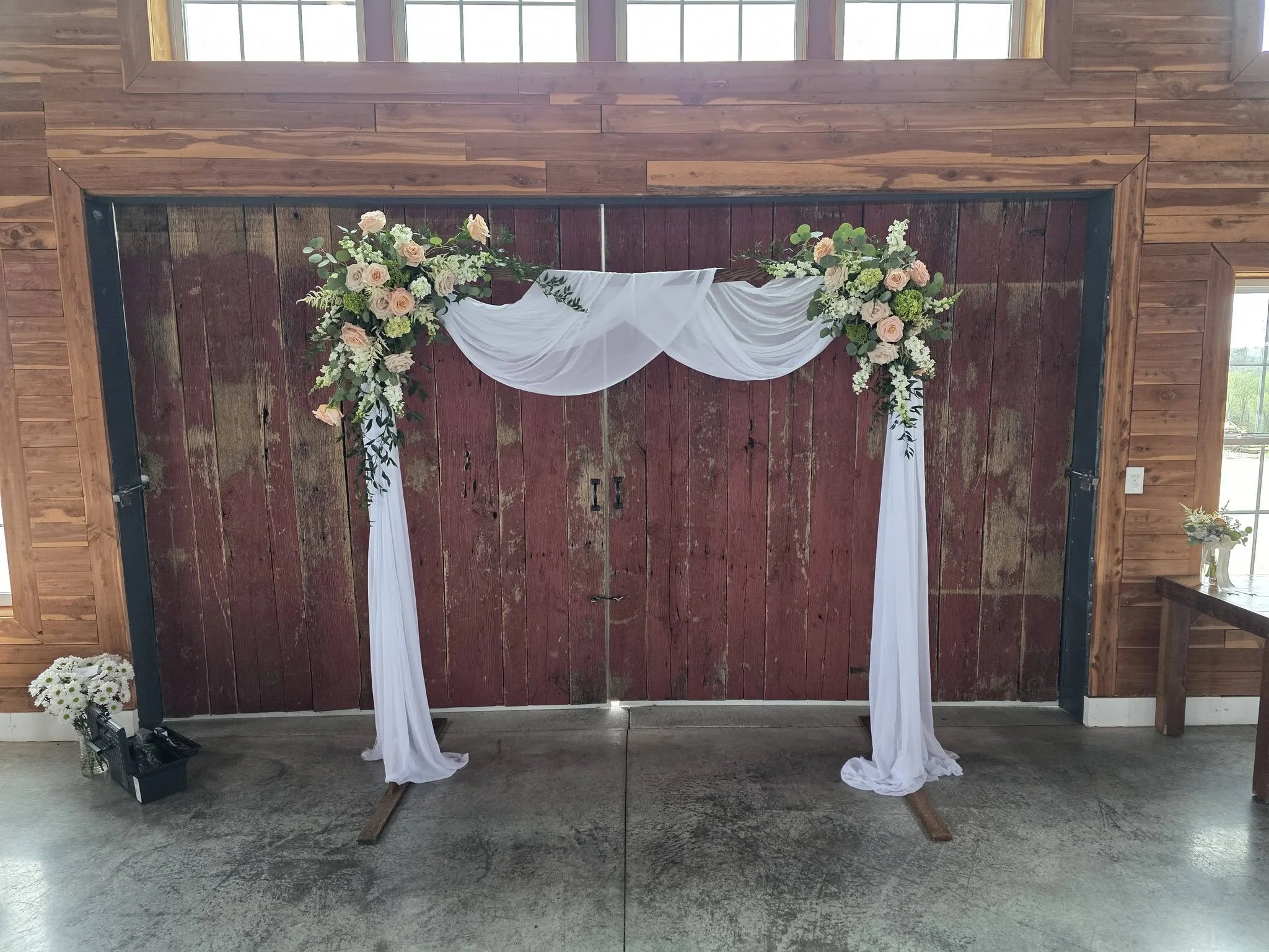 A wedding arch decorated with white sheer fabric and floral arrangements, set against a rustic red barn door. To the left is a small bouquet of white flowers in a jar, and to the right, a table or stand is partly visible.