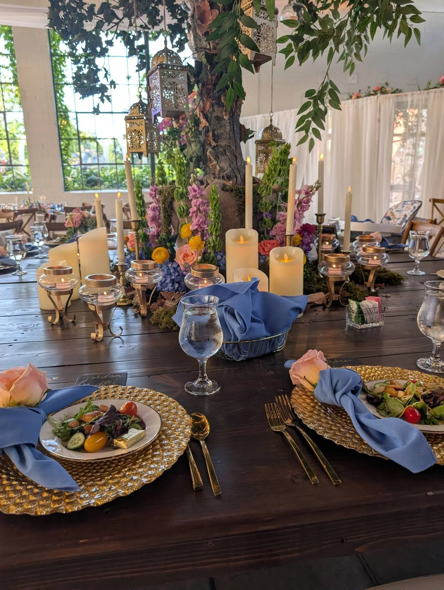 A decorated dining table with floral centerpieces, candles, and elegant tableware in a bright, airy room with large windows and greenery outside.