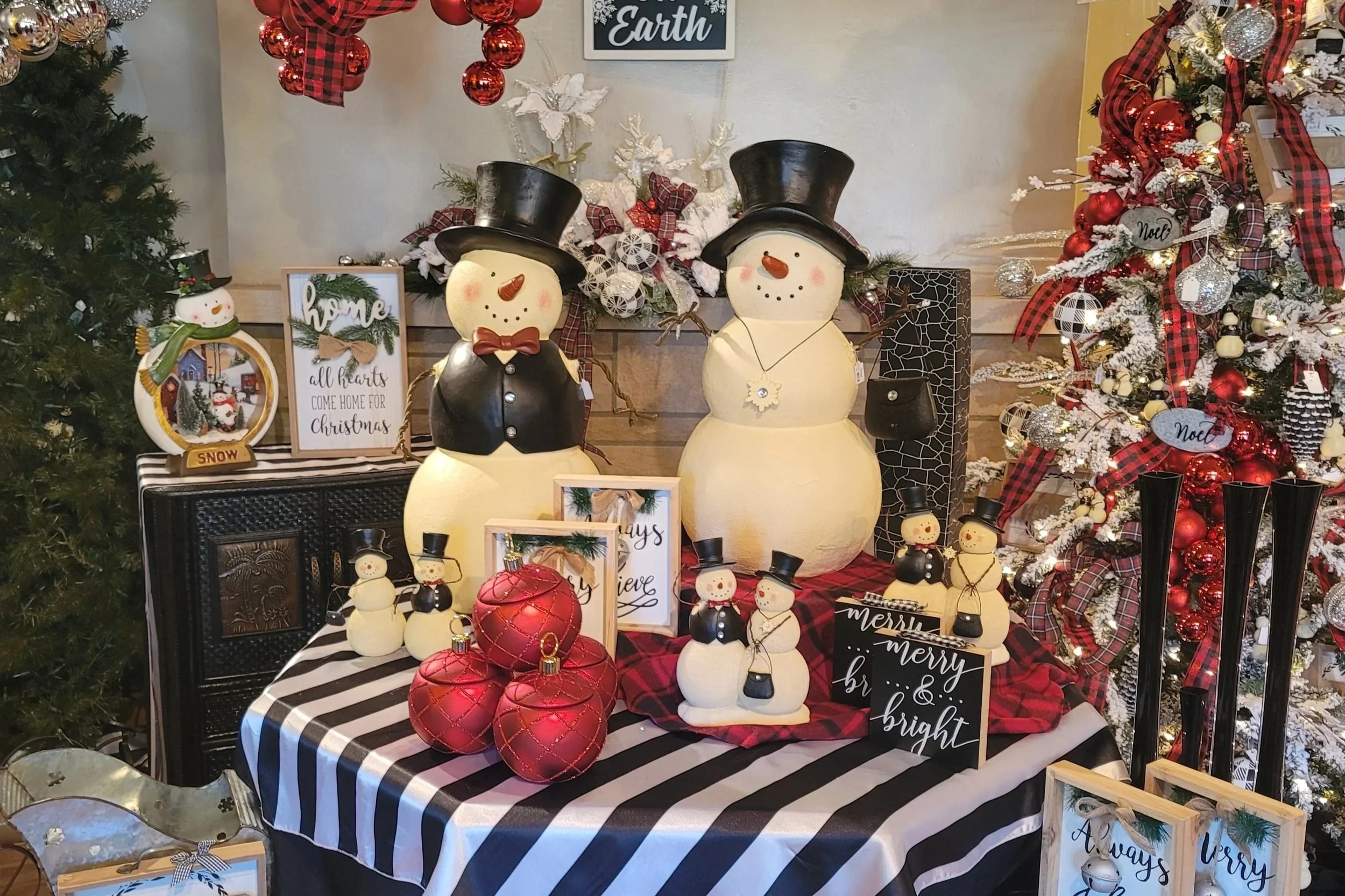Decorative Christmas display featuring two snowman figures with black top hats, one with a red bow tie and the other with a white snowflake necklace, surrounded by red ornaments, snowman figurines, framed signs with holiday messages, and a snowy Christmas tree decorated with red and white ornaments and ribbons. The setup is on a table covered with a black and white striped tablecloth.