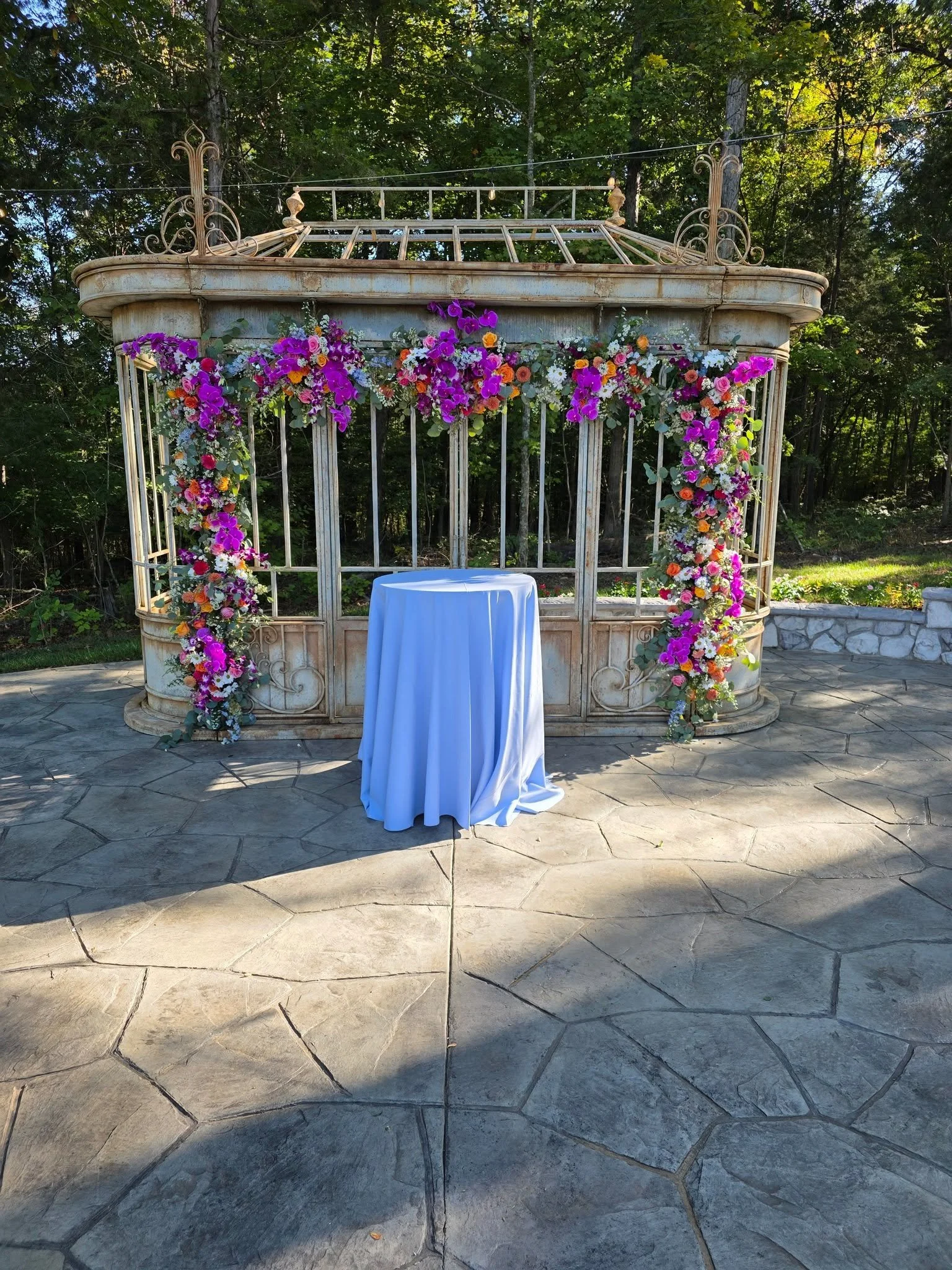 An outdoor setting with a white table covered with a white cloth in front of a decorated metal gazebo-style structure adorned with vibrant multicolored flowers. The background features lush green trees, and the ground is paved with large stone tiles.