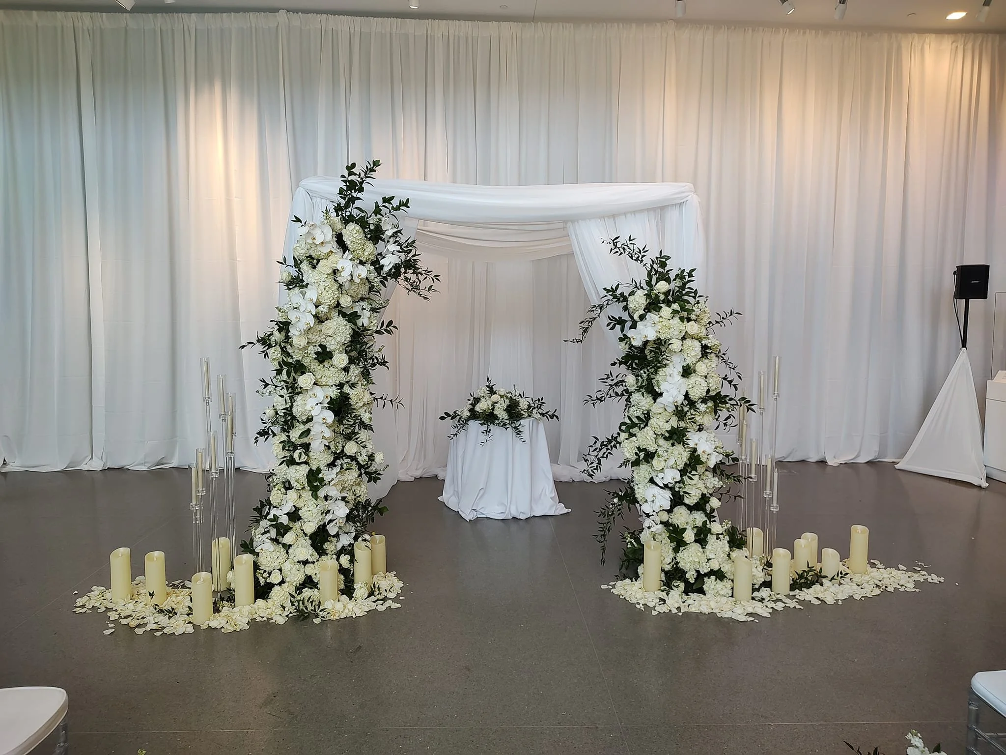 White floral wedding arch with candles at the base, set against a white curtain backdrop.