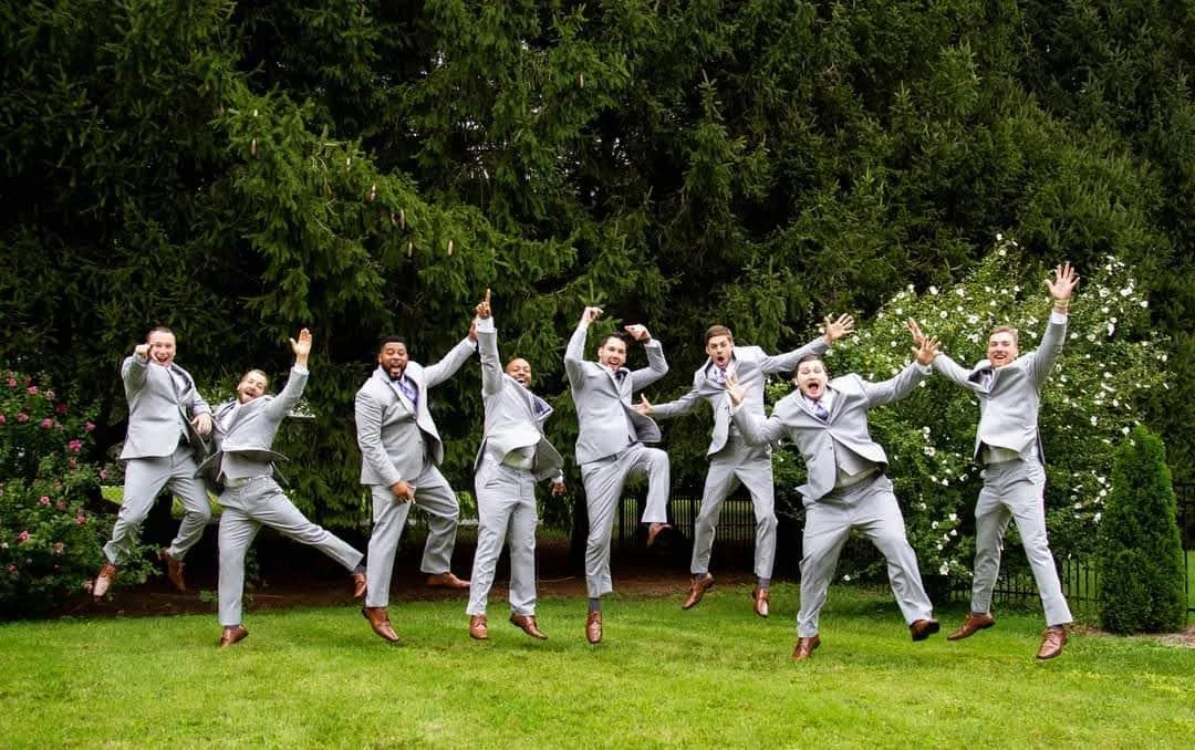 Group of eight men in gray suits jumping in the air outdoors in front of trees and bushes.