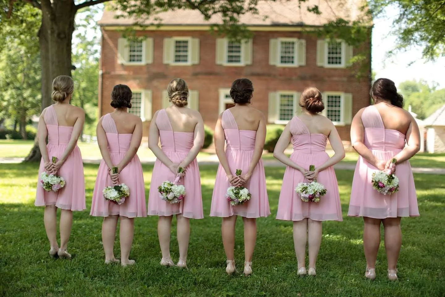 Six women in pink dresses holding bouquets, standing outdoors on grass with a large brick house and trees in the background.
