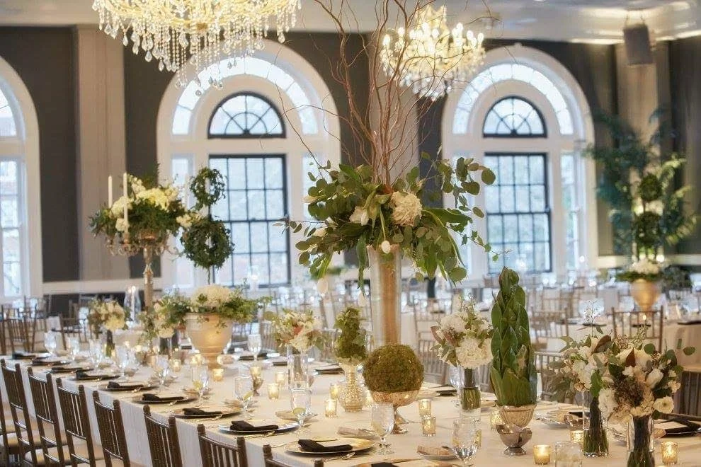Elegant banquet hall decorated for a wedding or event with floral centerpieces, candles, chandeliers, and large arched windows.