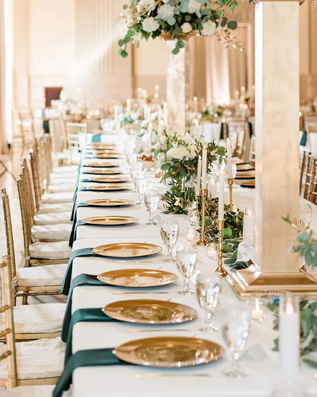 Elegant banquet table decorated with candles, floral arrangements, gold chargers, wine glasses, and white tablecloths in a well-lit room.