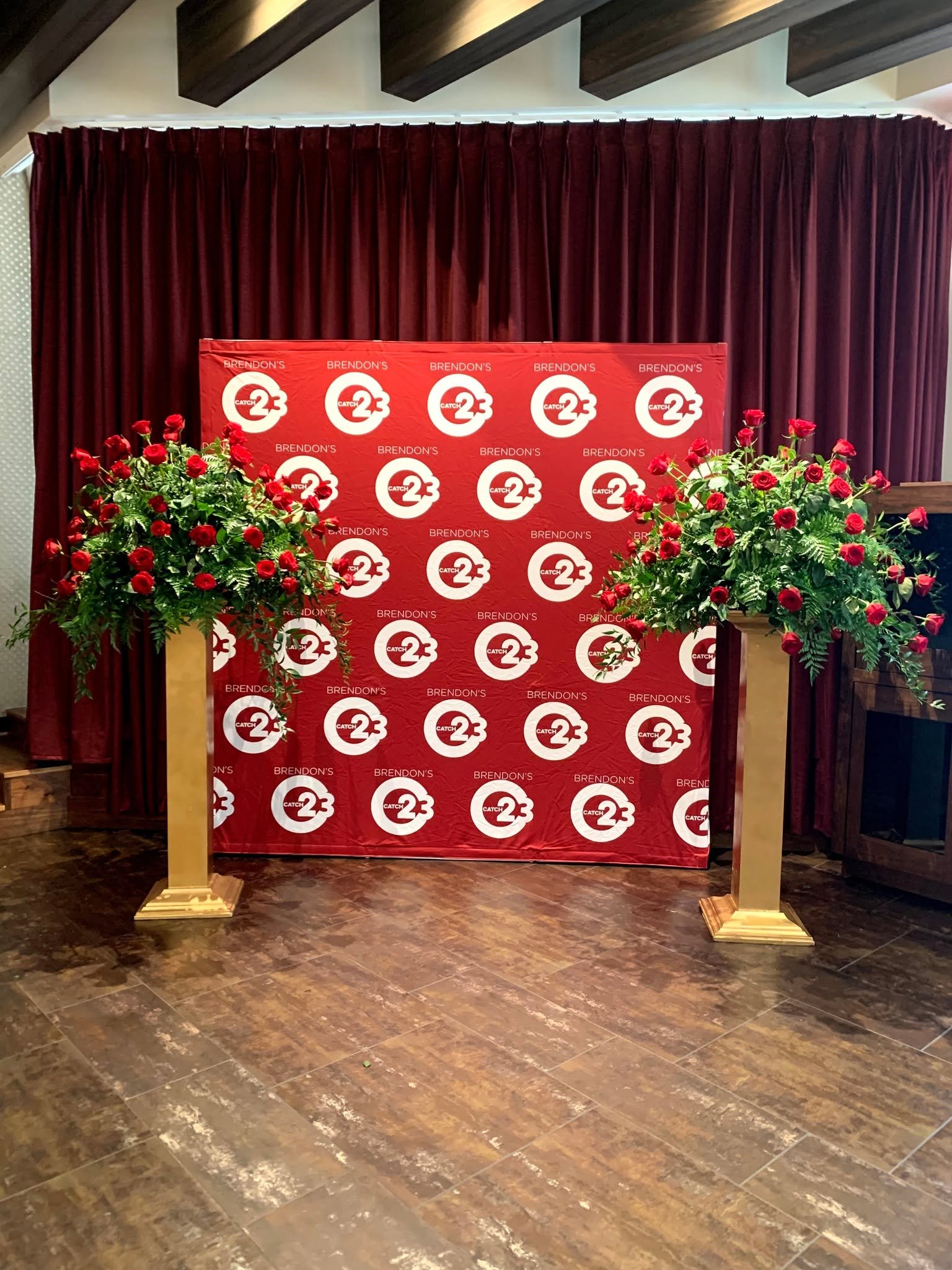 A decorative backdrop with red fabric curtains and a branded step and repeat banner featuring the logo for 'Catch 23' and 'Brendon's', flanked by two large flower arrangements of red roses and green foliage on golden stands, located on a wooden floor.