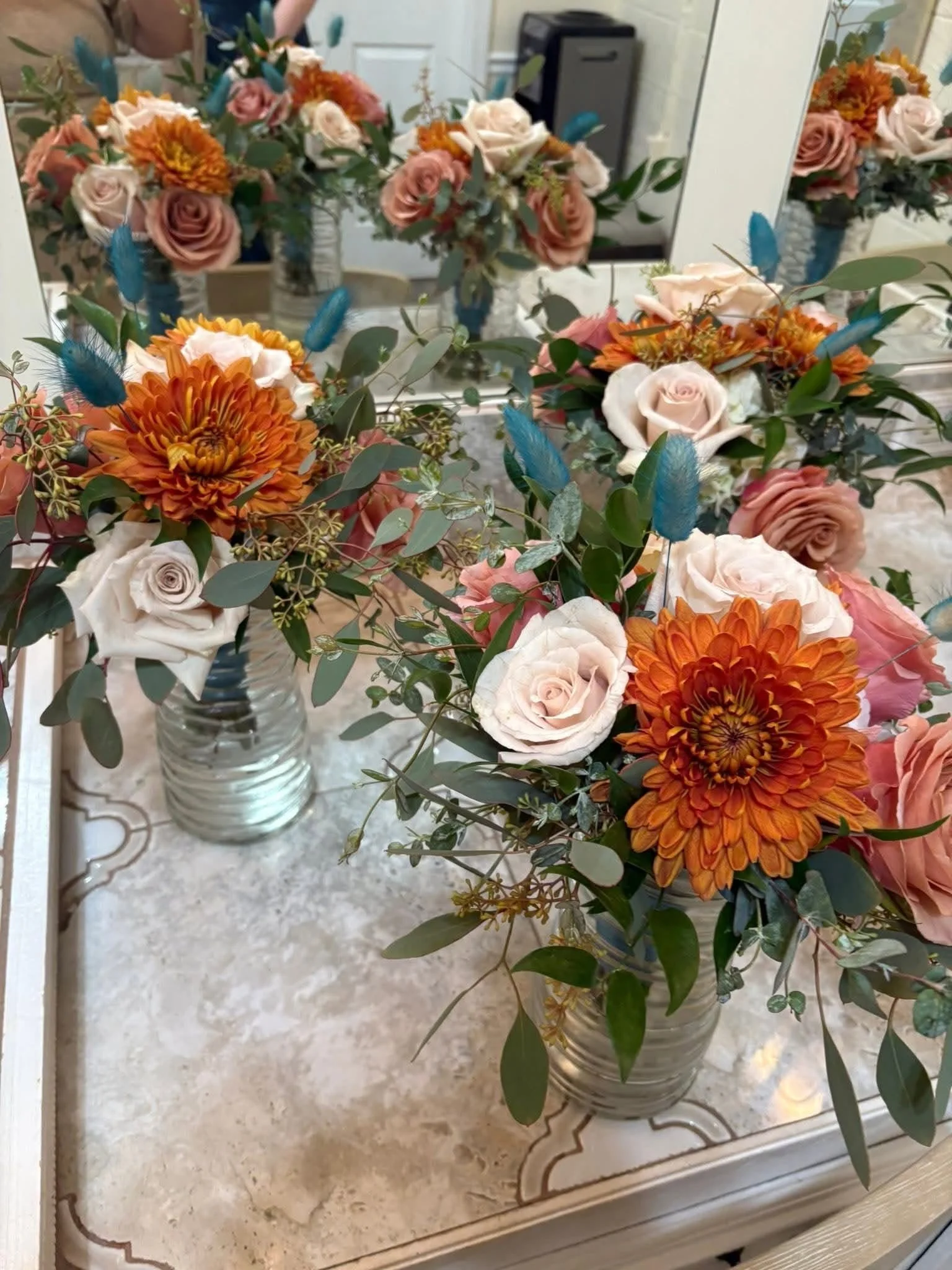 Three vases with peach, white, and orange flowers, placed on a mirror table with a marble surface, surrounded by eucalyptus leaves and decorative blue feathers, with a mirror reflecting the floral arrangements.