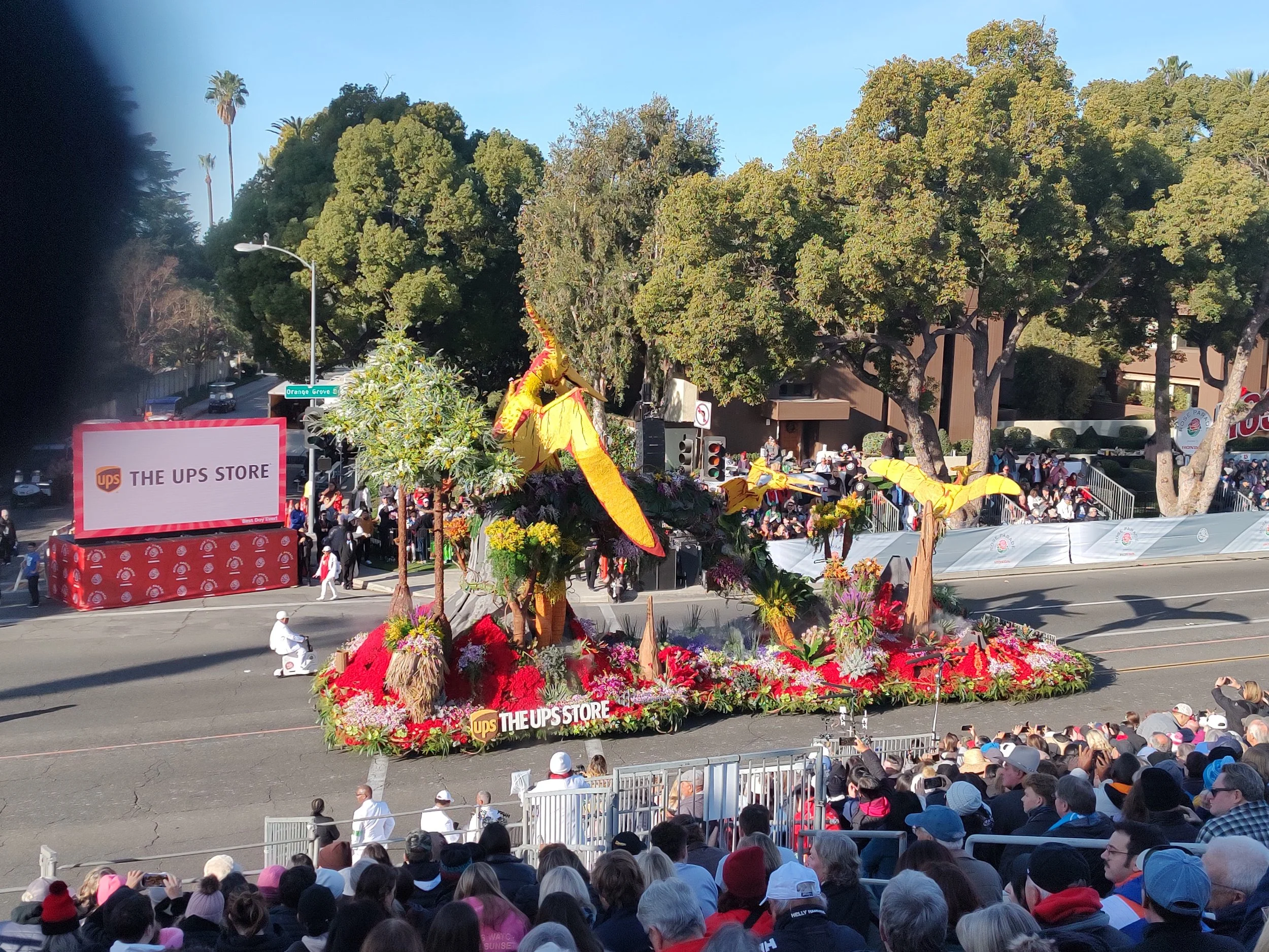 A colorful parade float with a large dragon made of flowers and plants, surrounded by a crowd of spectators in outdoor seating. The float features bright yellow, red, and green decorations and a banner for The UPS Store.