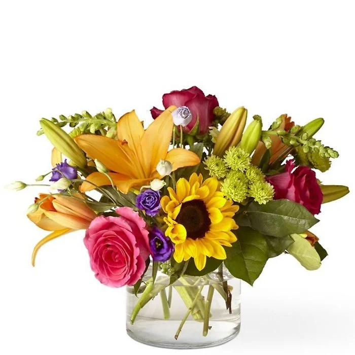 Colorful flower bouquet with roses, lilies, sunflowers, and other flowers in a clear glass vase against a white background.