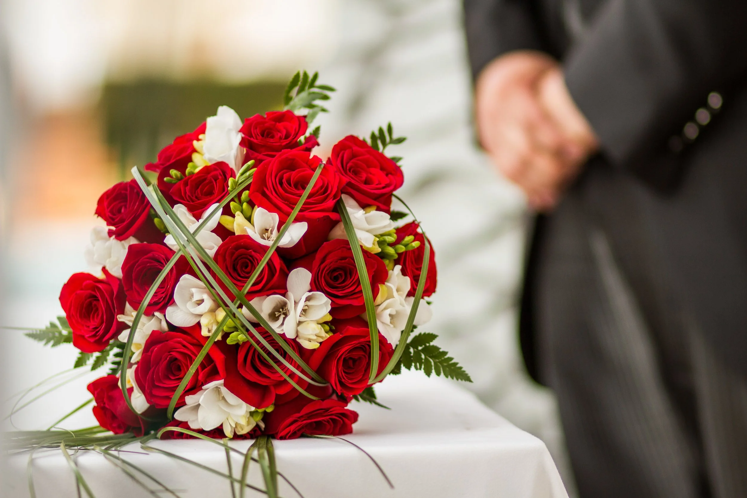 A close-up of a bridal bouquet with red roses, white orchids, and green foliage, held by a person dressed in a suit.
