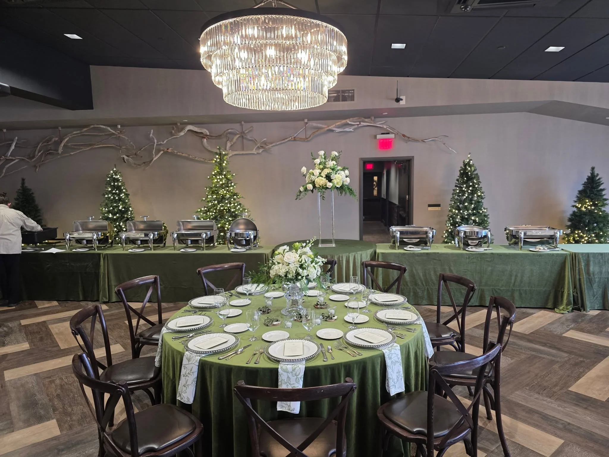 A banquet hall decorated for a holiday celebration with Christmas trees adorned with lights, a large floral centerpiece, and a round table set with place settings, plates, glasses, and silverware.