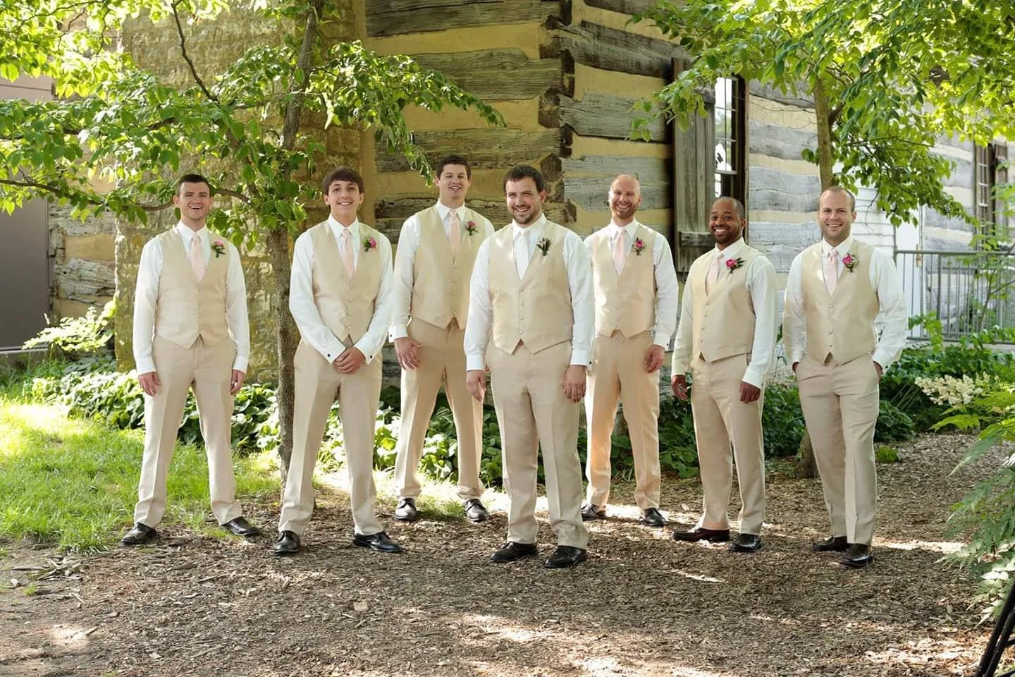 Group of eight groomsmens in cream-colored suits with pink ties, standing outdoors under a tree in front of a rustic wooden building, smiling.